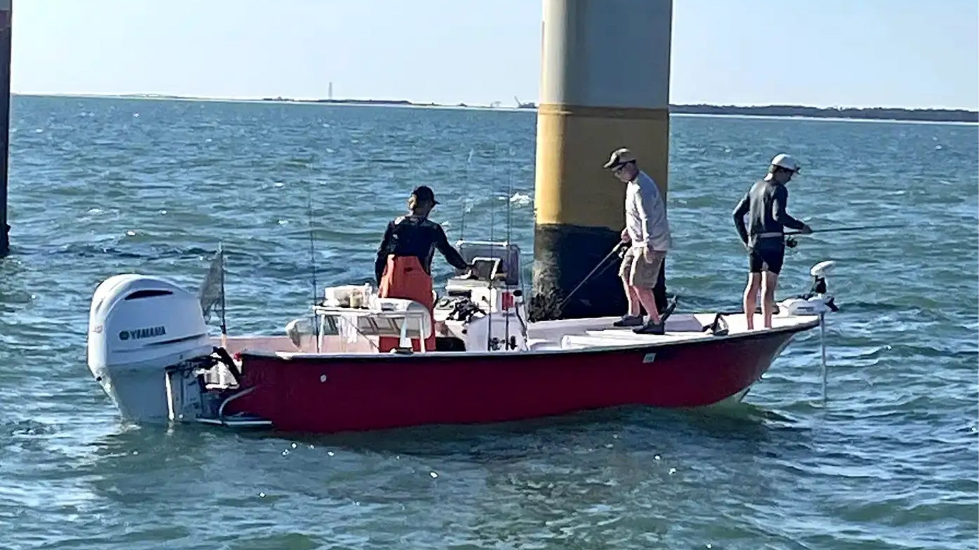 Three people fishing from a red boat docked beside a large metal piling in the water.