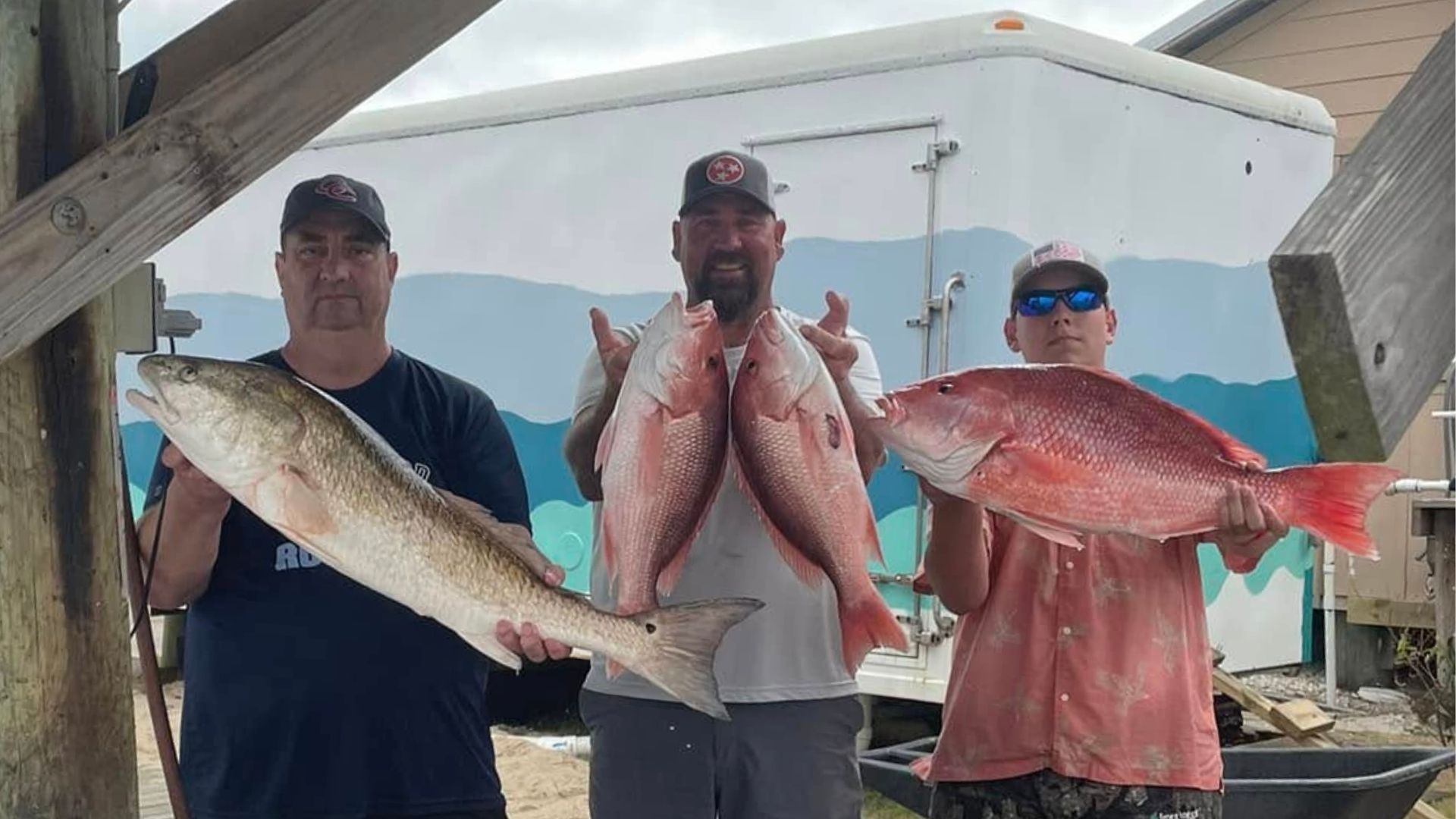 Three people hold up large fish in front of a docked boat, posing for a catch photo.