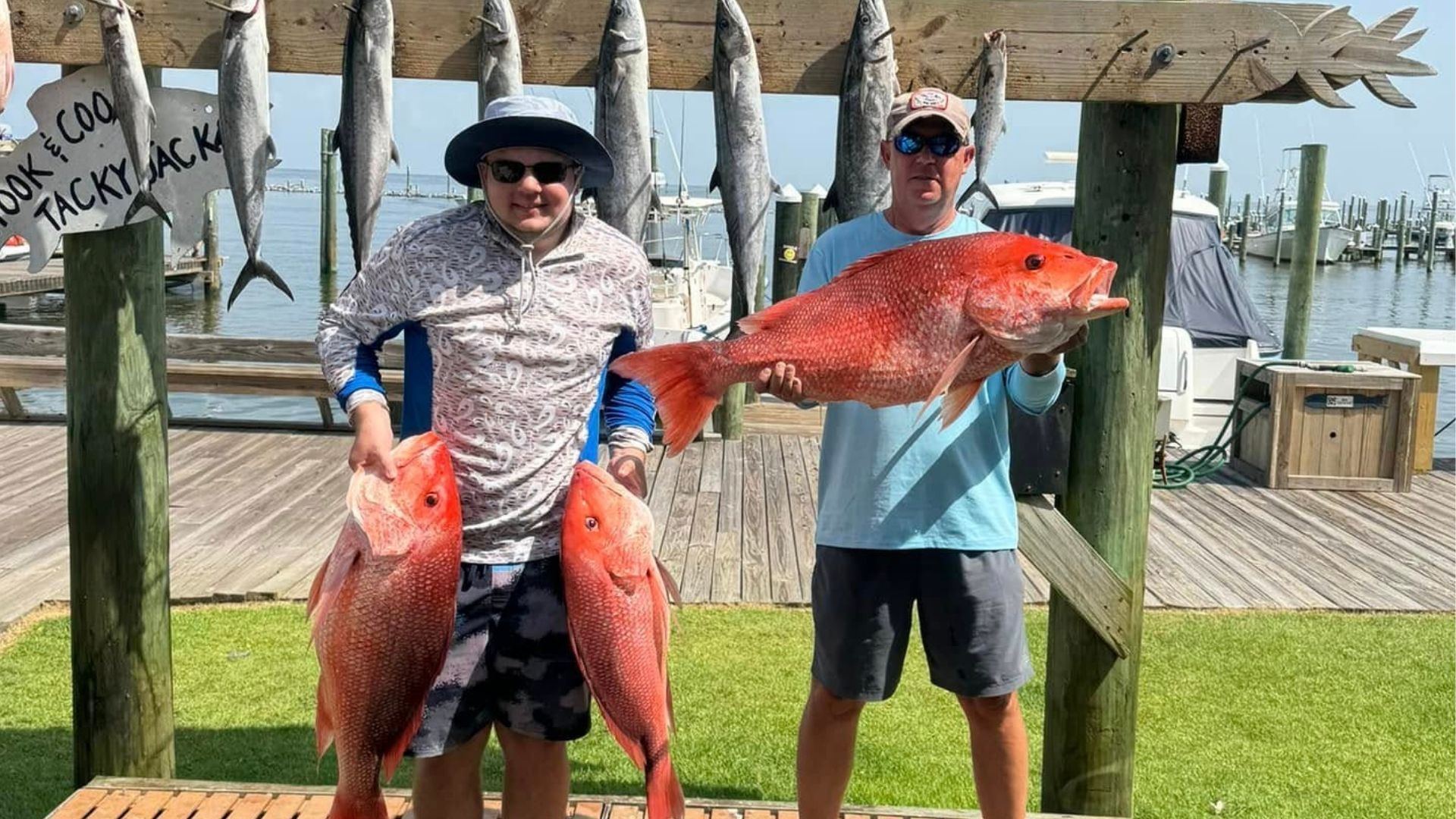 Two people holding large red fish on a dock by the water, with another person in the background.