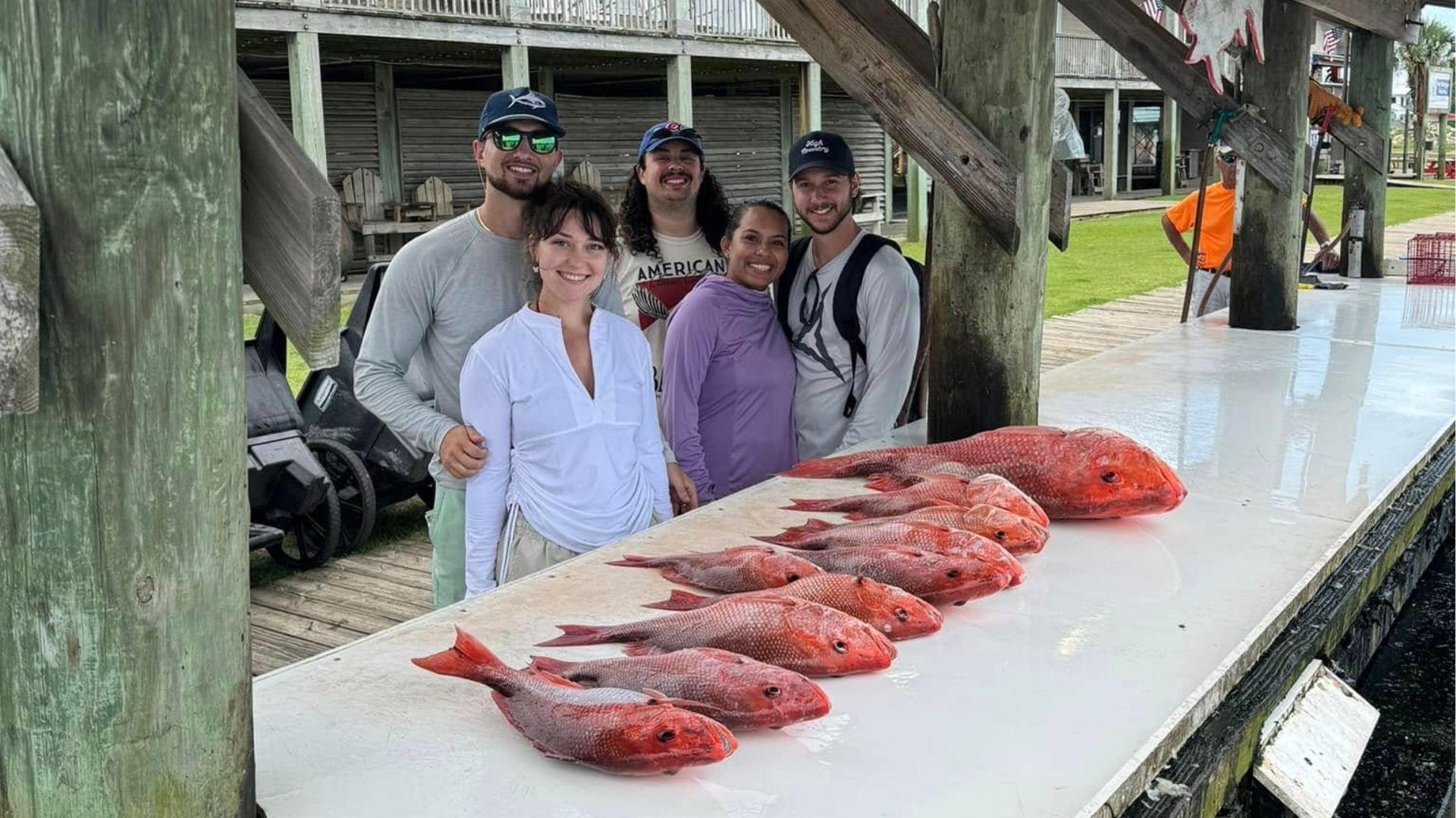 People standing beside a long table of freshly caught red fish at a dock under a wooden shelter