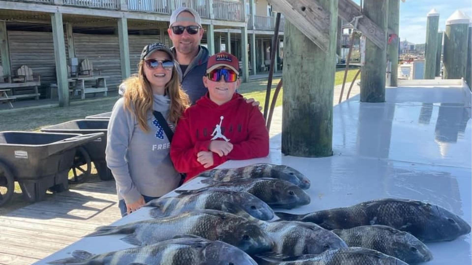 Three people posing with a pile of fish on a dock, wearing sunglasses in bright sunlight