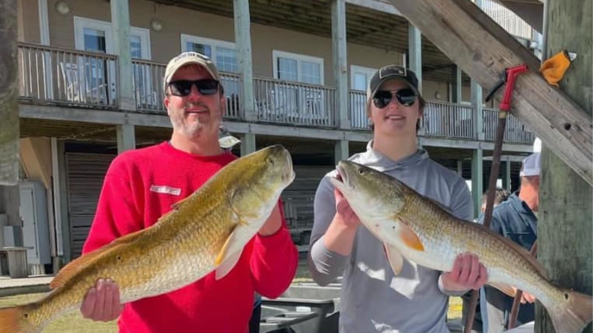 Two men holding large fish on a dock, smiling in front of a marina building.