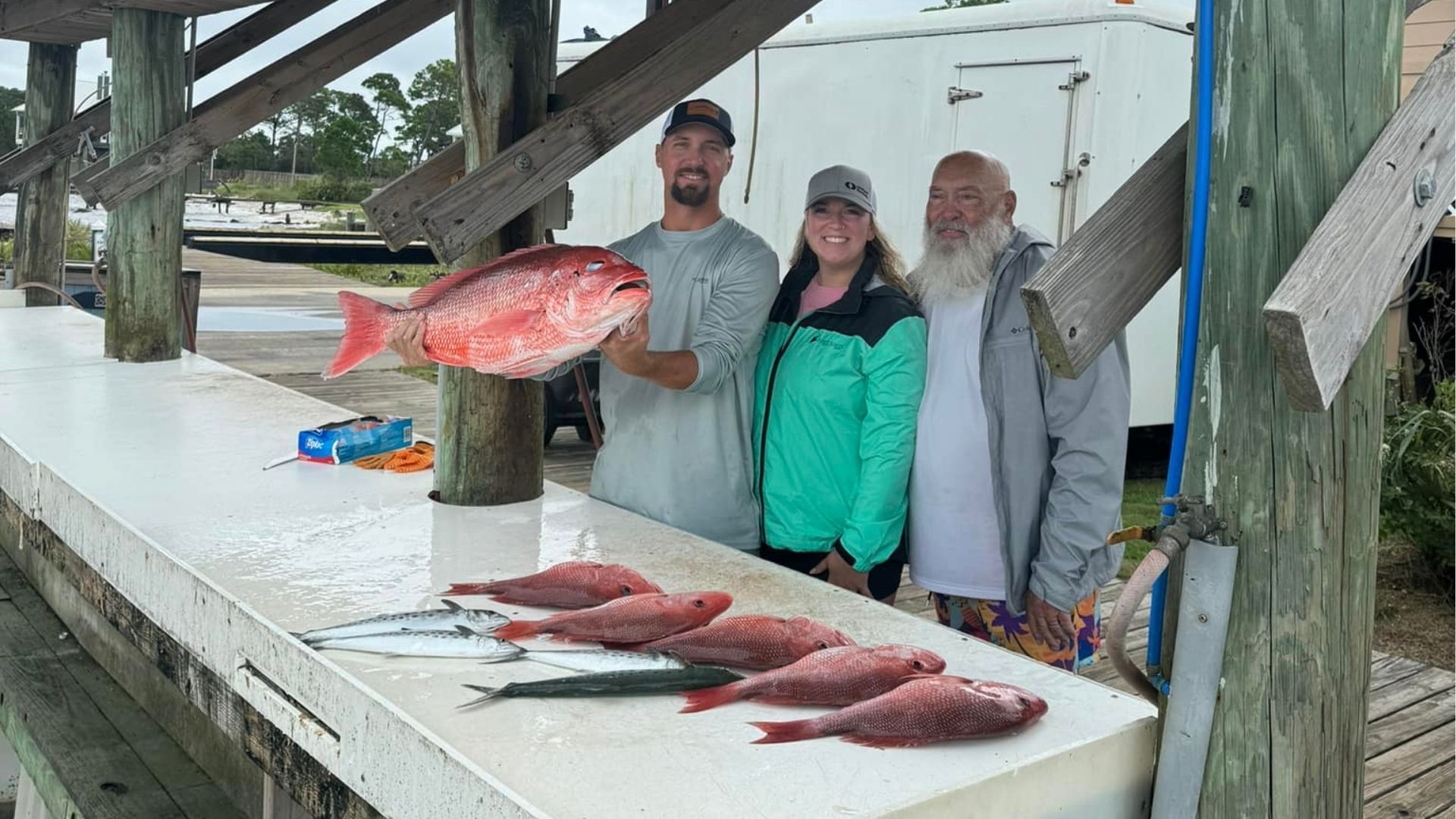 People posing on a dock beside several large fish laid out on a table under a wooden shelter.