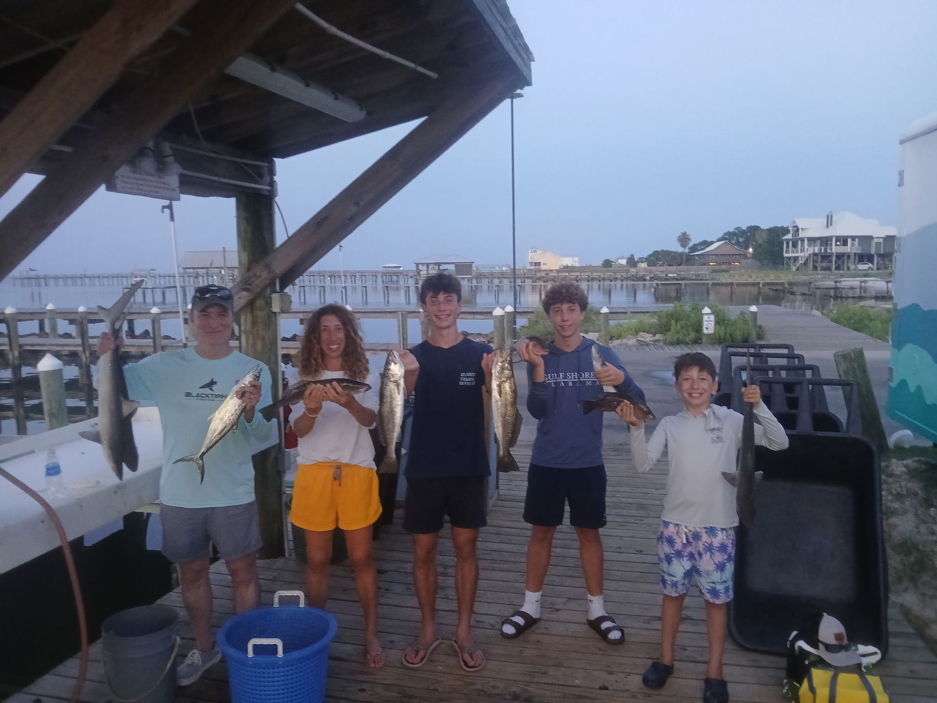 Five people pose on a dock holding fish, with a boat and calm water in the background.