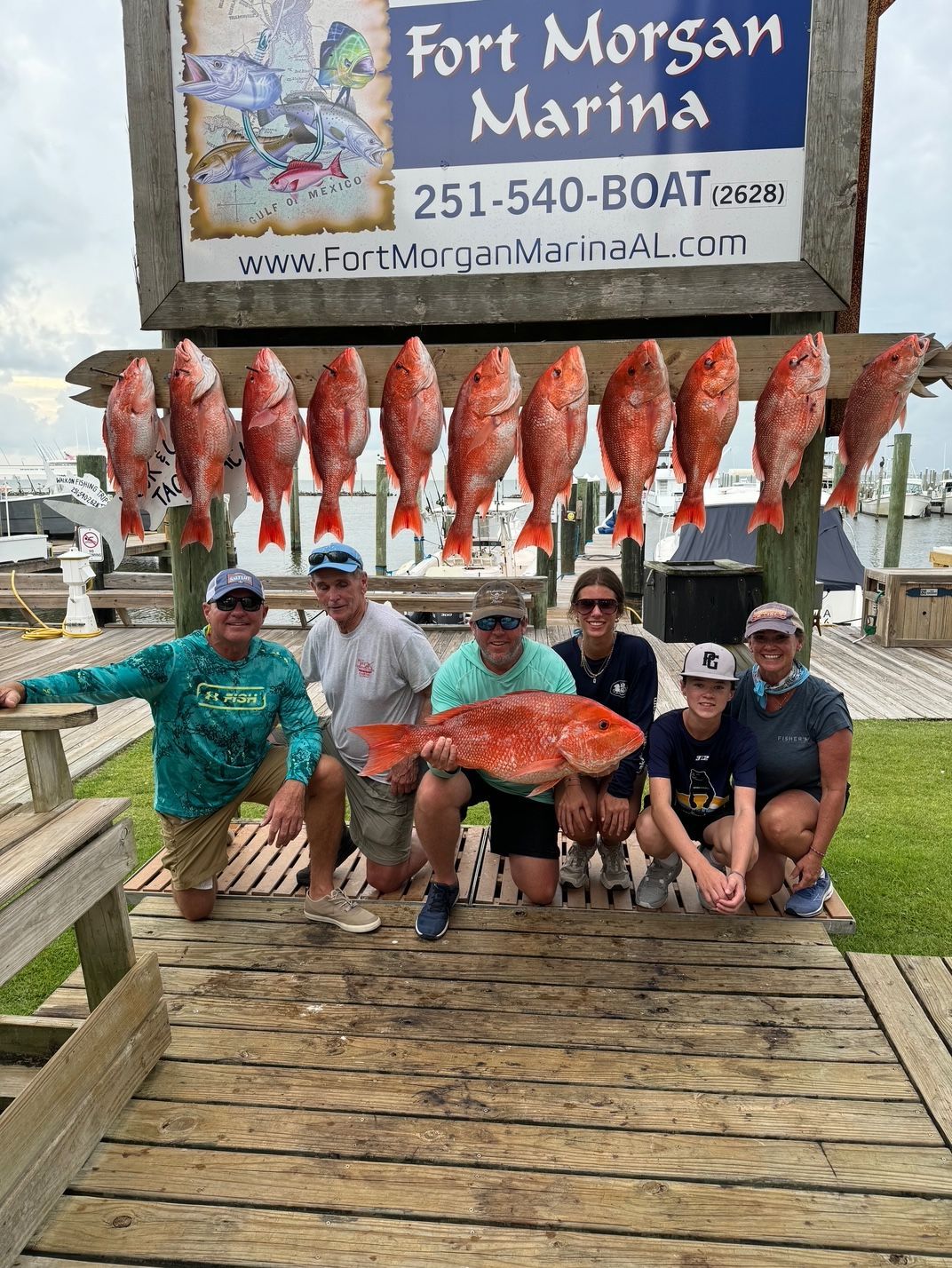 Six fishermen pose with large red snapper in front of Fort Morgan Marina sign and hanging fish display.