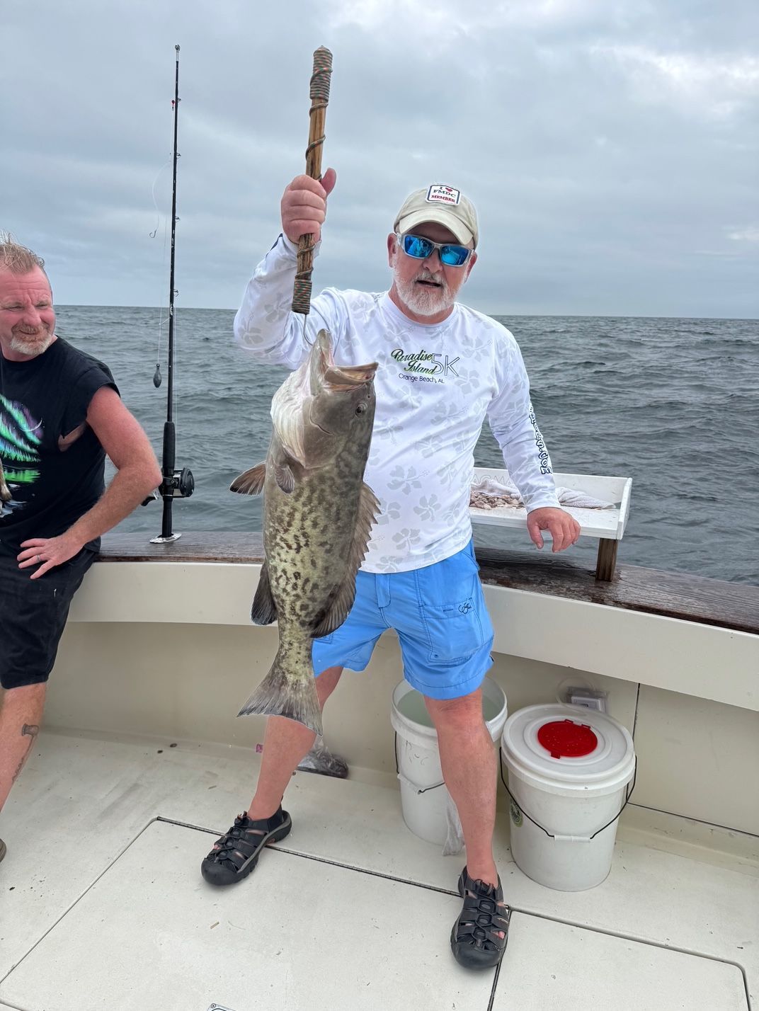 Person on a boat proudly holds up a large spotted fish by the lake under cloudy skies