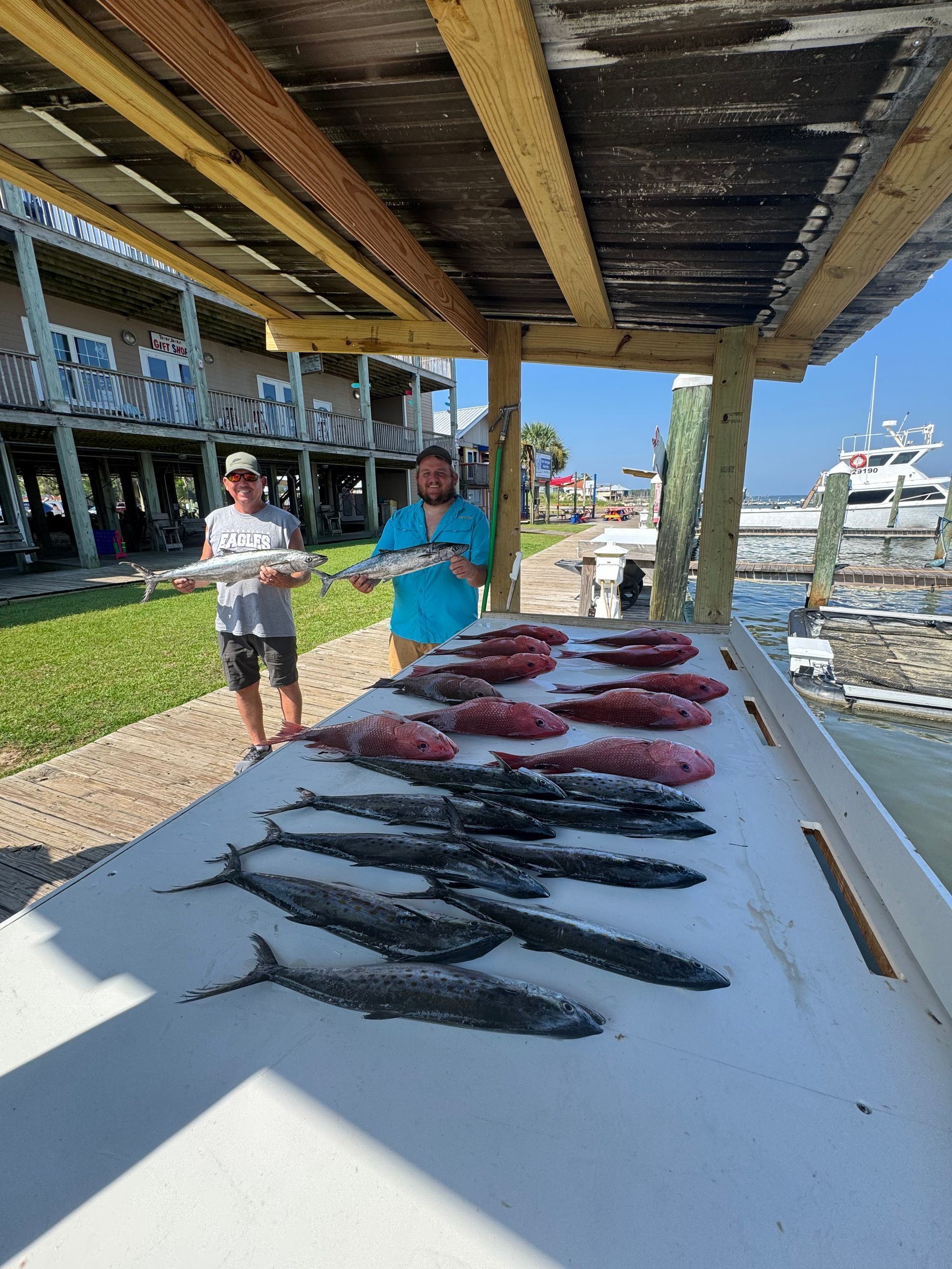 Fresh fish laid out on a table at a dock, with two people standing nearby by the water.
