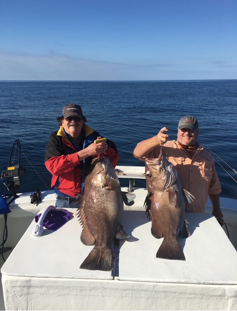 Two anglers on a boat hold up large fish against a blue ocean backdrop.
