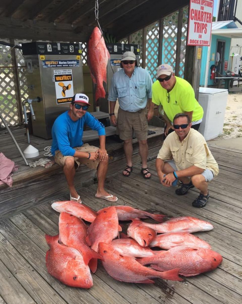 Four people pose beside a large pile of red fish on a wooden dock at a marina.