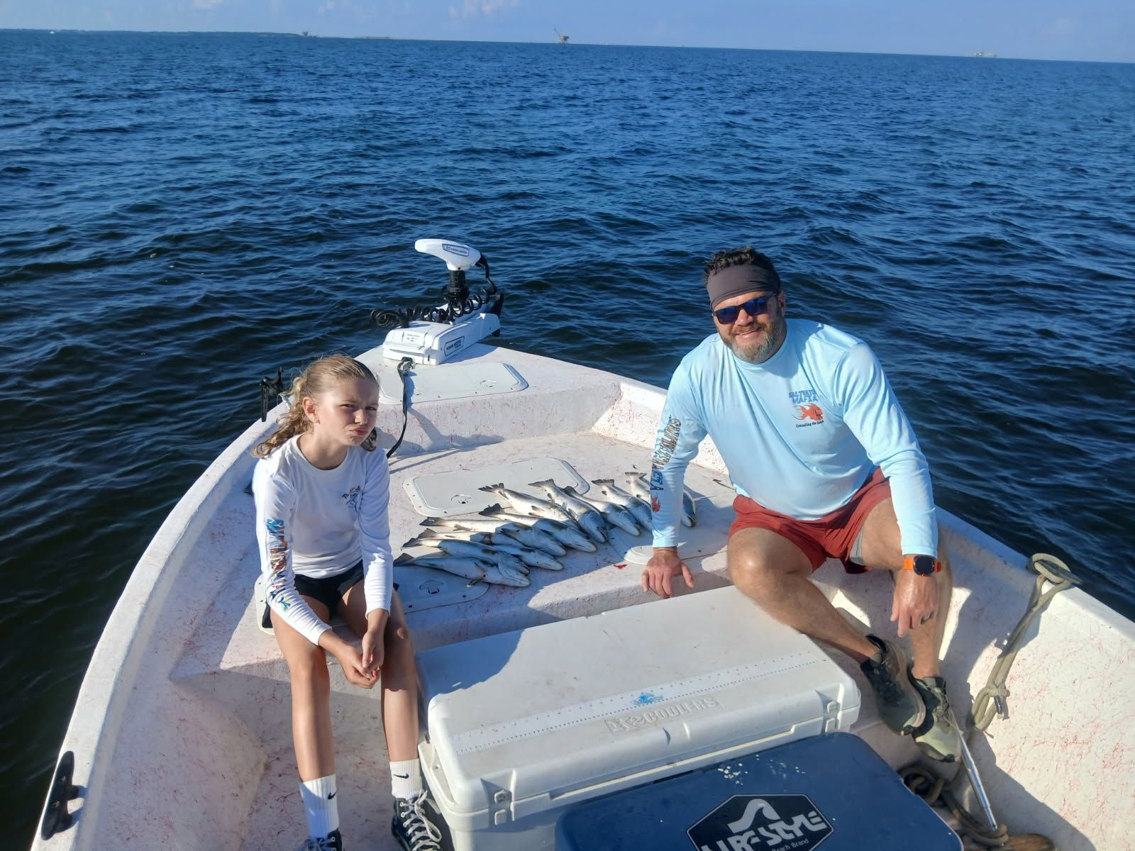Two people seated on the bow of a boat on open water, with bright blue sea behind them.