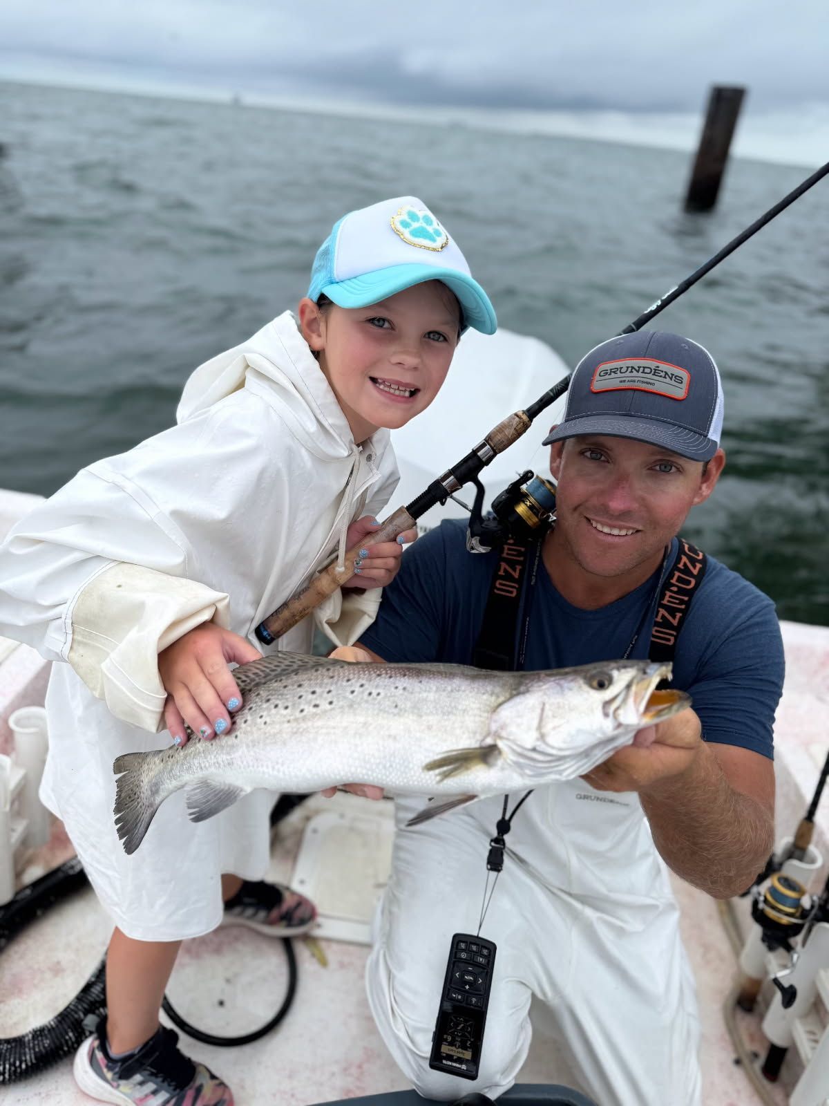 Two people on a boat holding a large fish, wearing caps and rain gear on a cloudy day