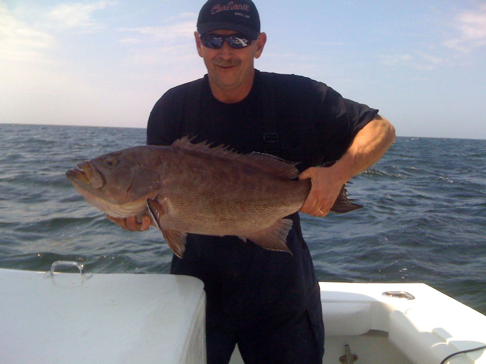 A person wearing a dark shirt and sunglasses holds a large brown fish aboard a boat on the water.