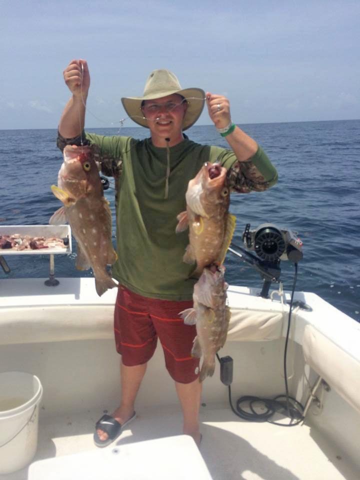A person wearing a sun hat stands on a boat, holding three spotted fish over the ocean.