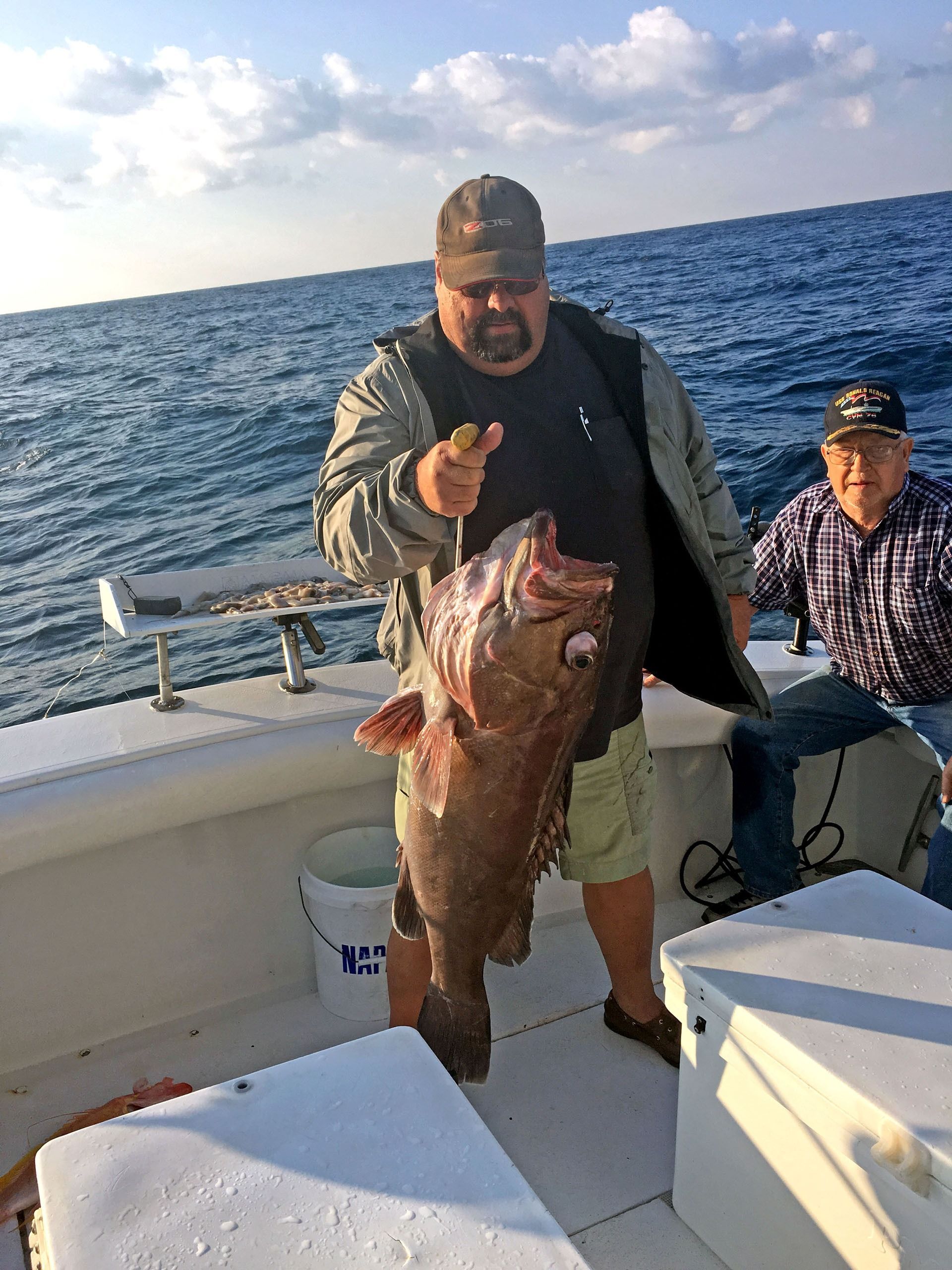 A person stands on a boat deck holding a large, dark fish, while another person sits nearby on the open water.