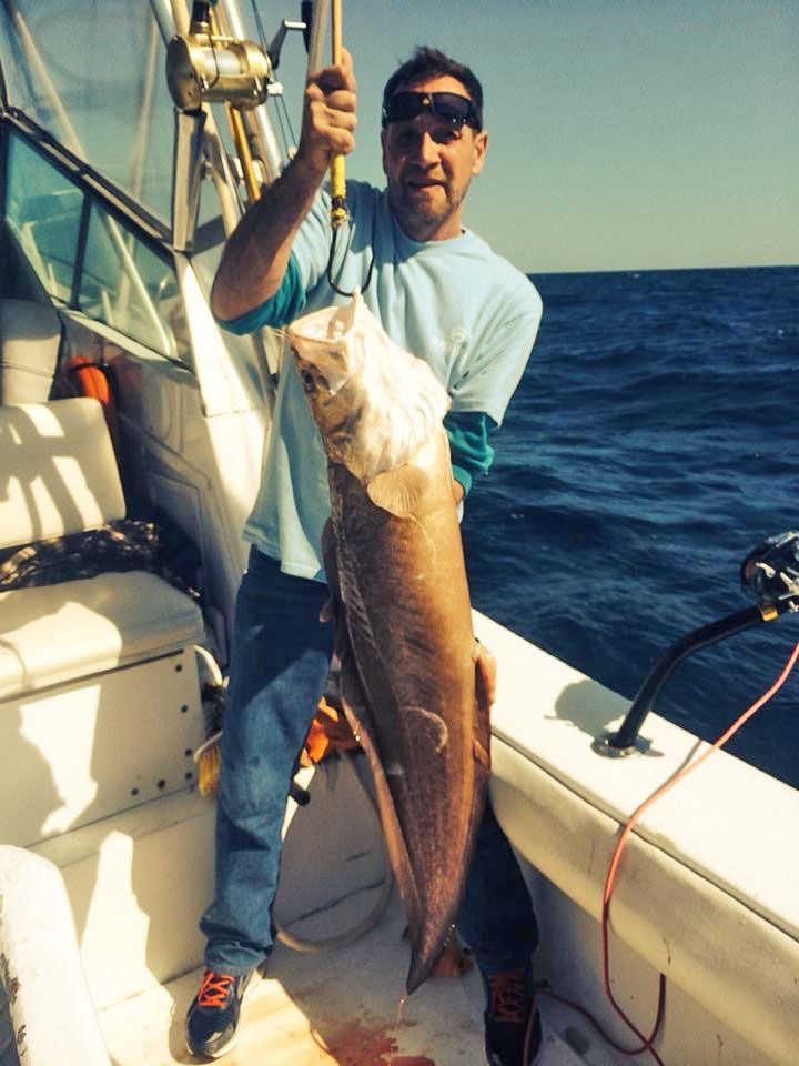 A person on a boat holding a large, freshly caught fish suspended from a scale.