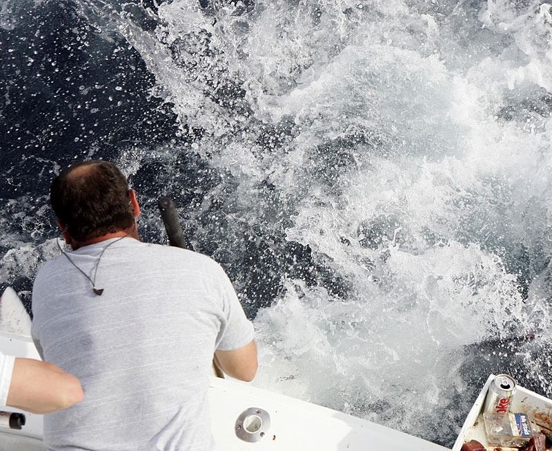 A person on a boat leans over the stern, watching water churn aggressively as a large fish breaches the surface.