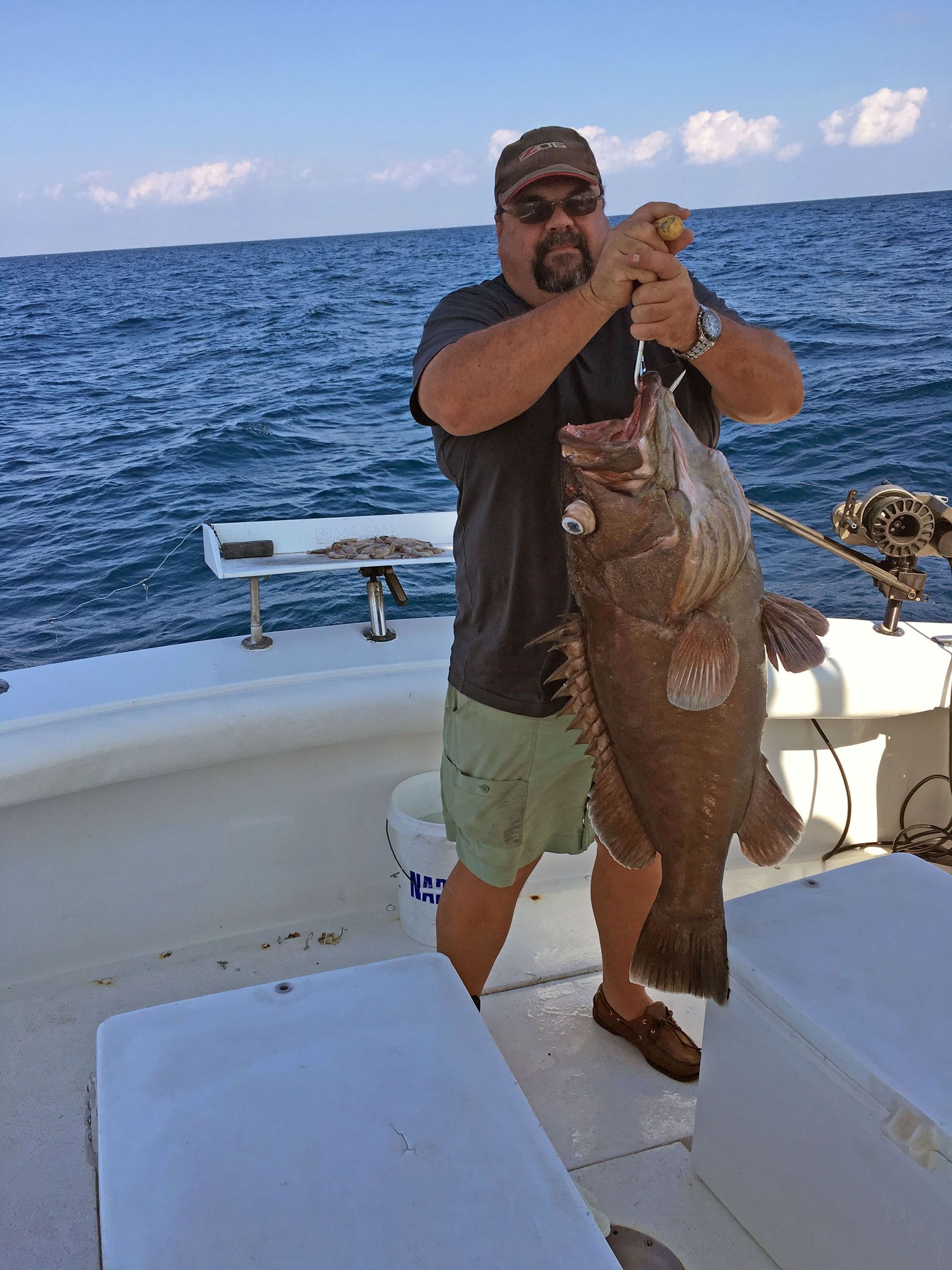 A person in a hat and shorts stands on a boat, holding a large fish caught from the open ocean.