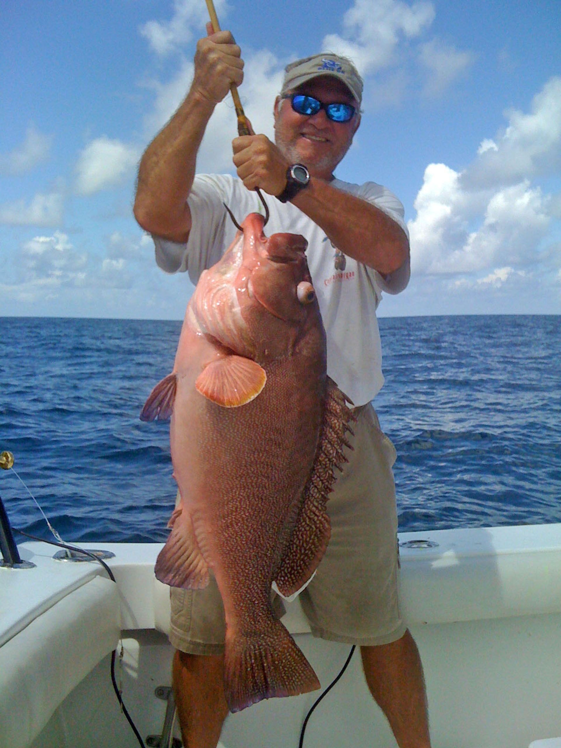 A person wearing sunglasses and a cap holds a large, reddish-brown fish by a hook on a boat under a sunny sky.