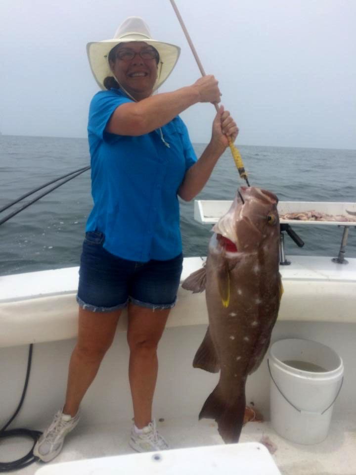 A person on a boat holds a gaff containing a large brown grouper with white spots against an ocean background.