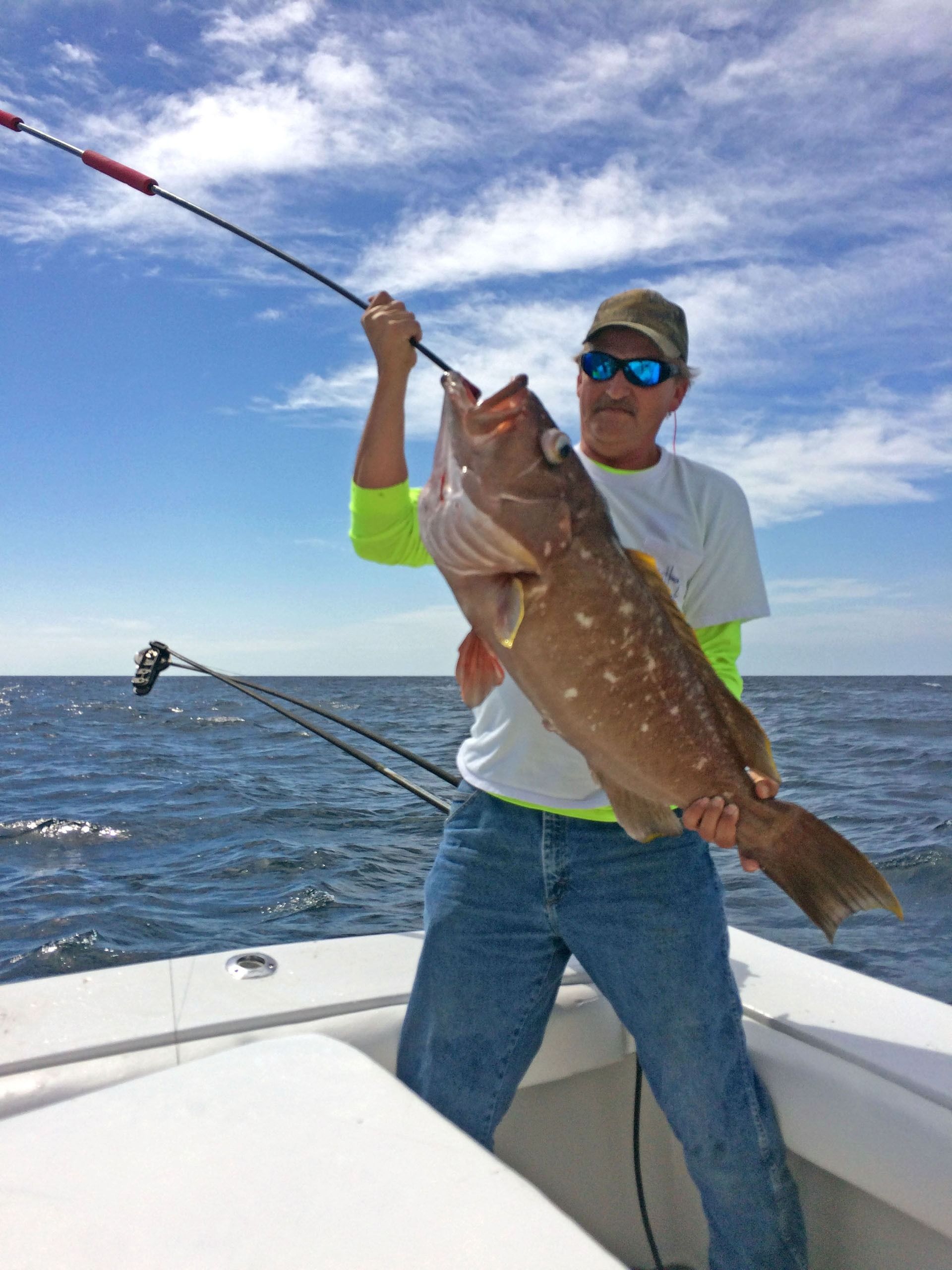 A person on a boat holds up a large grouper fish caught on a fishing line against a blue sky and ocean background.