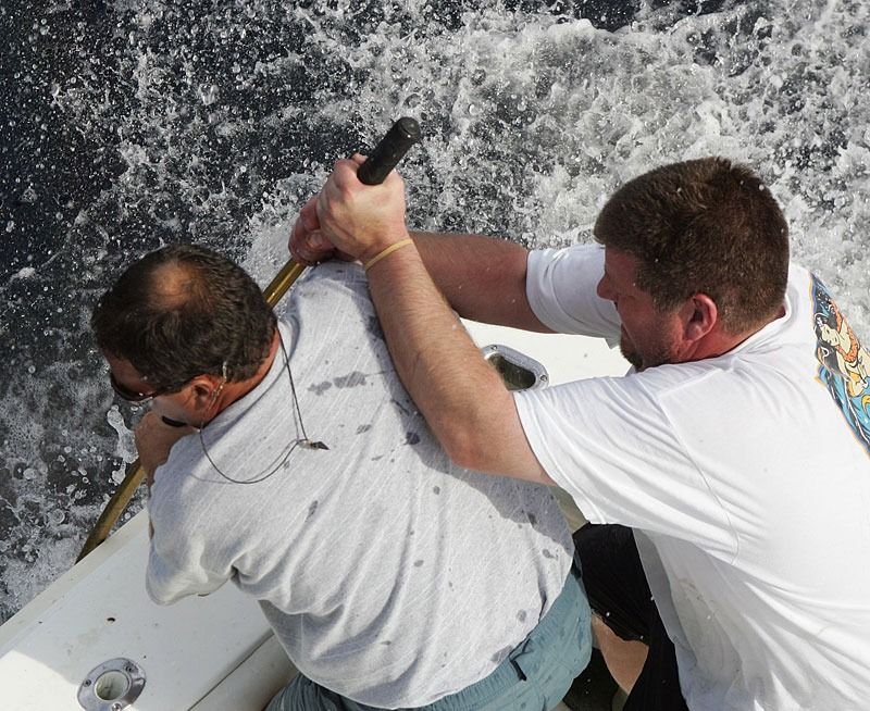 Two individuals on a boat using a long pole to reach into the water, with churning white water in the background.