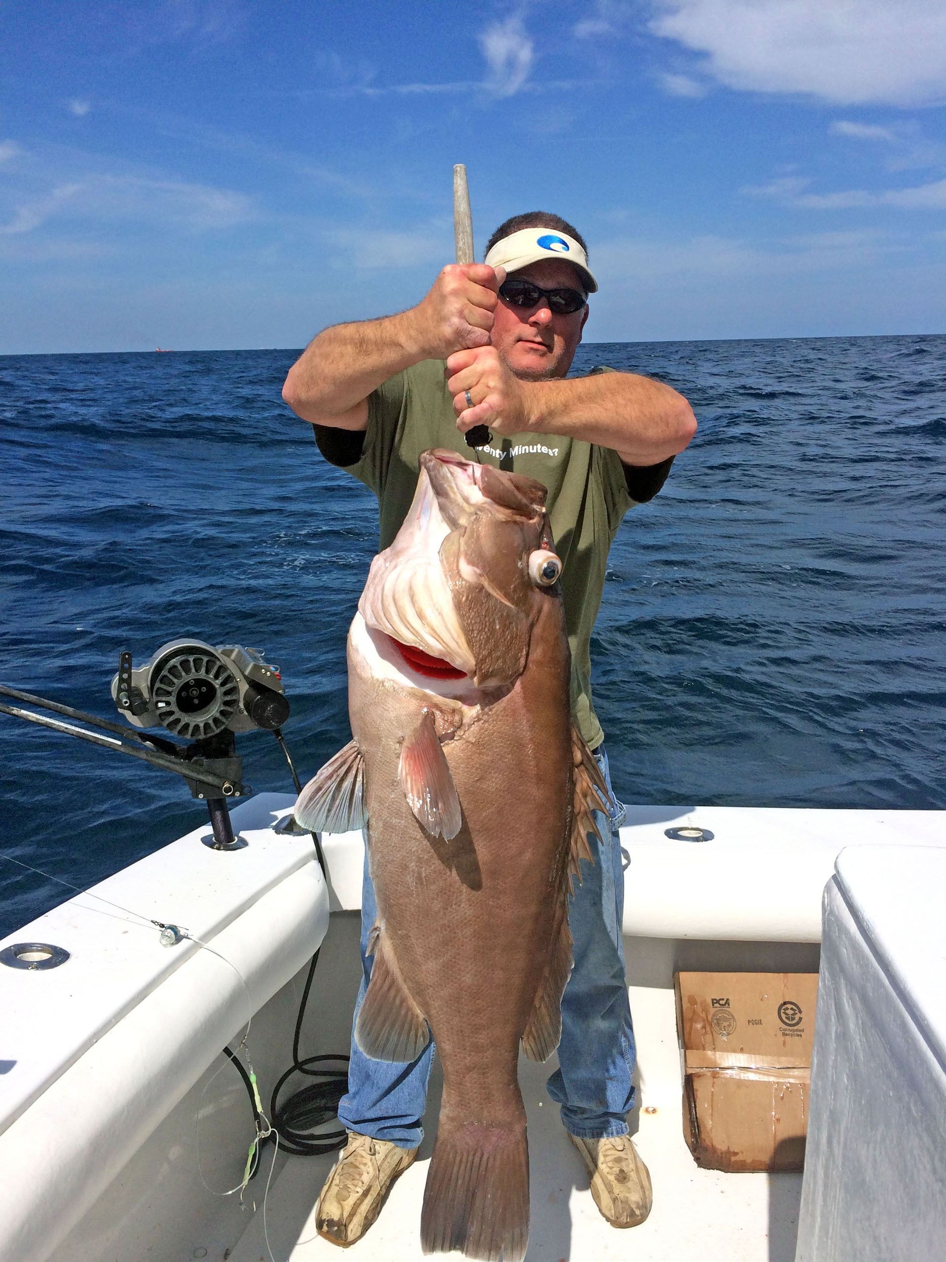 A person on a boat holding a large, brown grouper fish up by its mouth against a backdrop of the ocean.