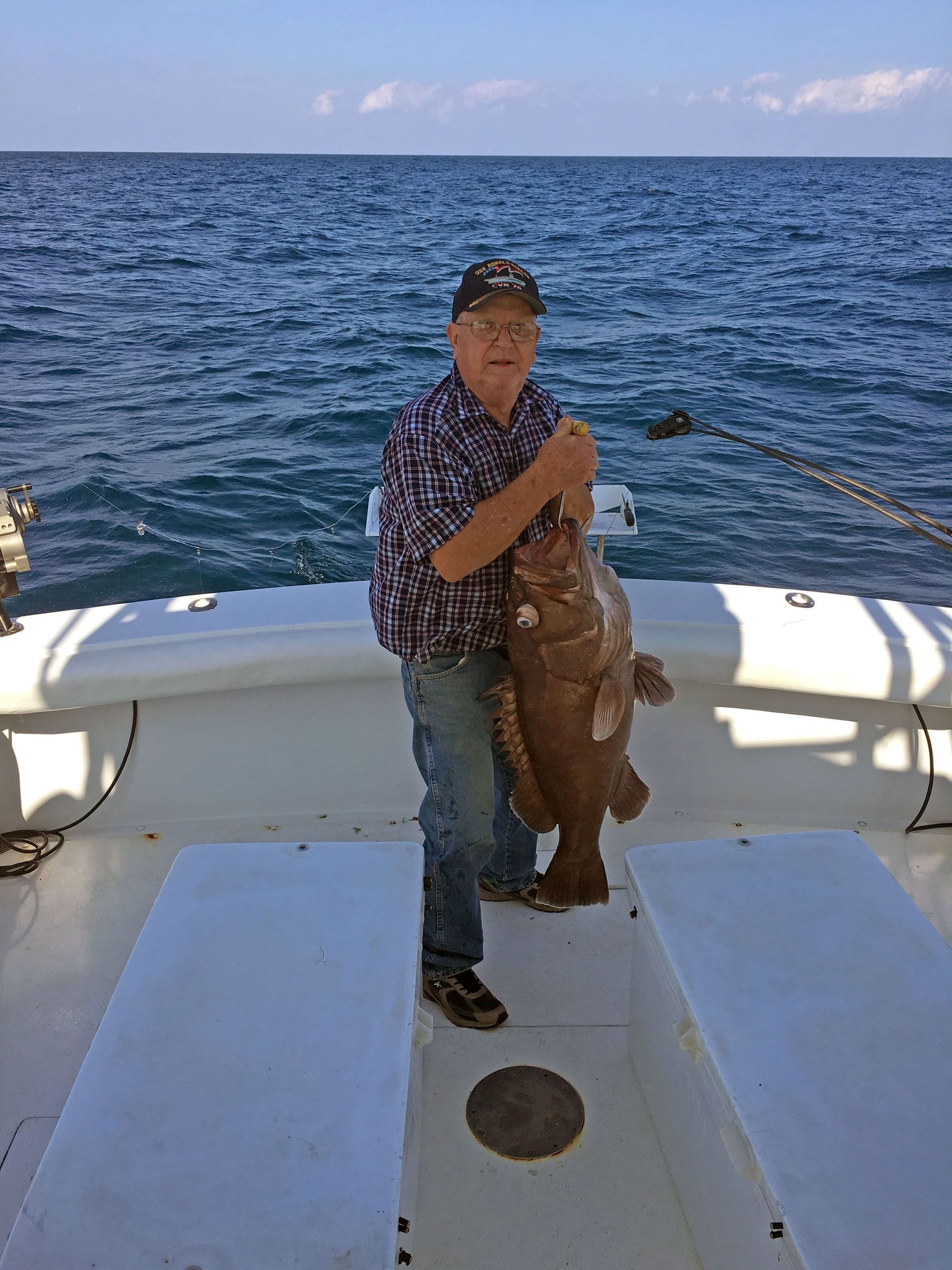 A person stands on a boat deck holding a large, brown fish over the ocean.