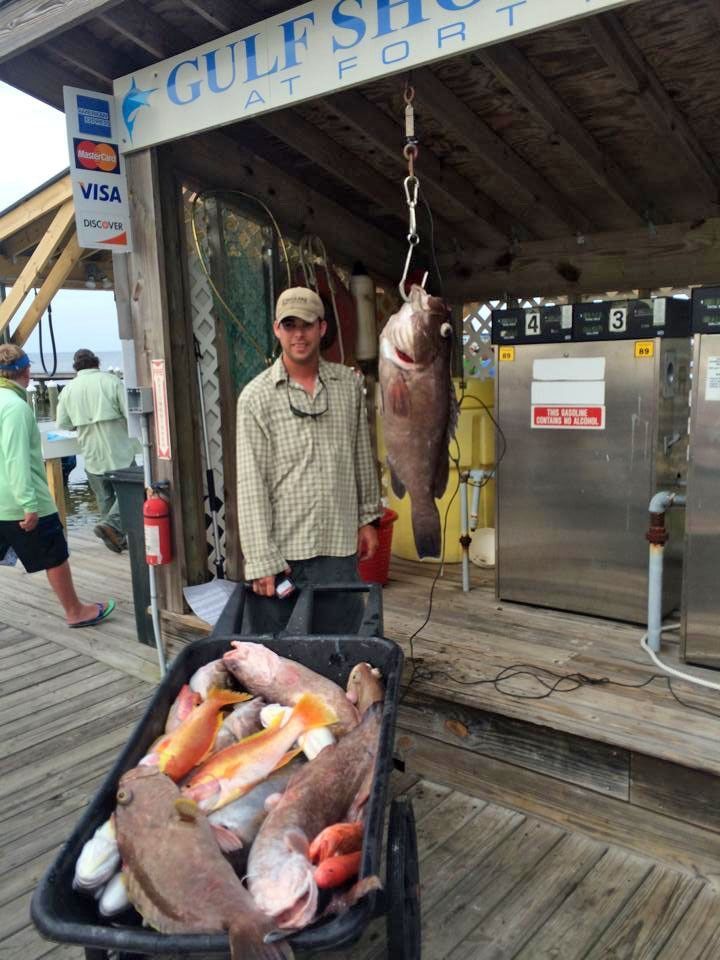 A person stands under a dock structure next to a cart full of fish and a large, hanging fish.
