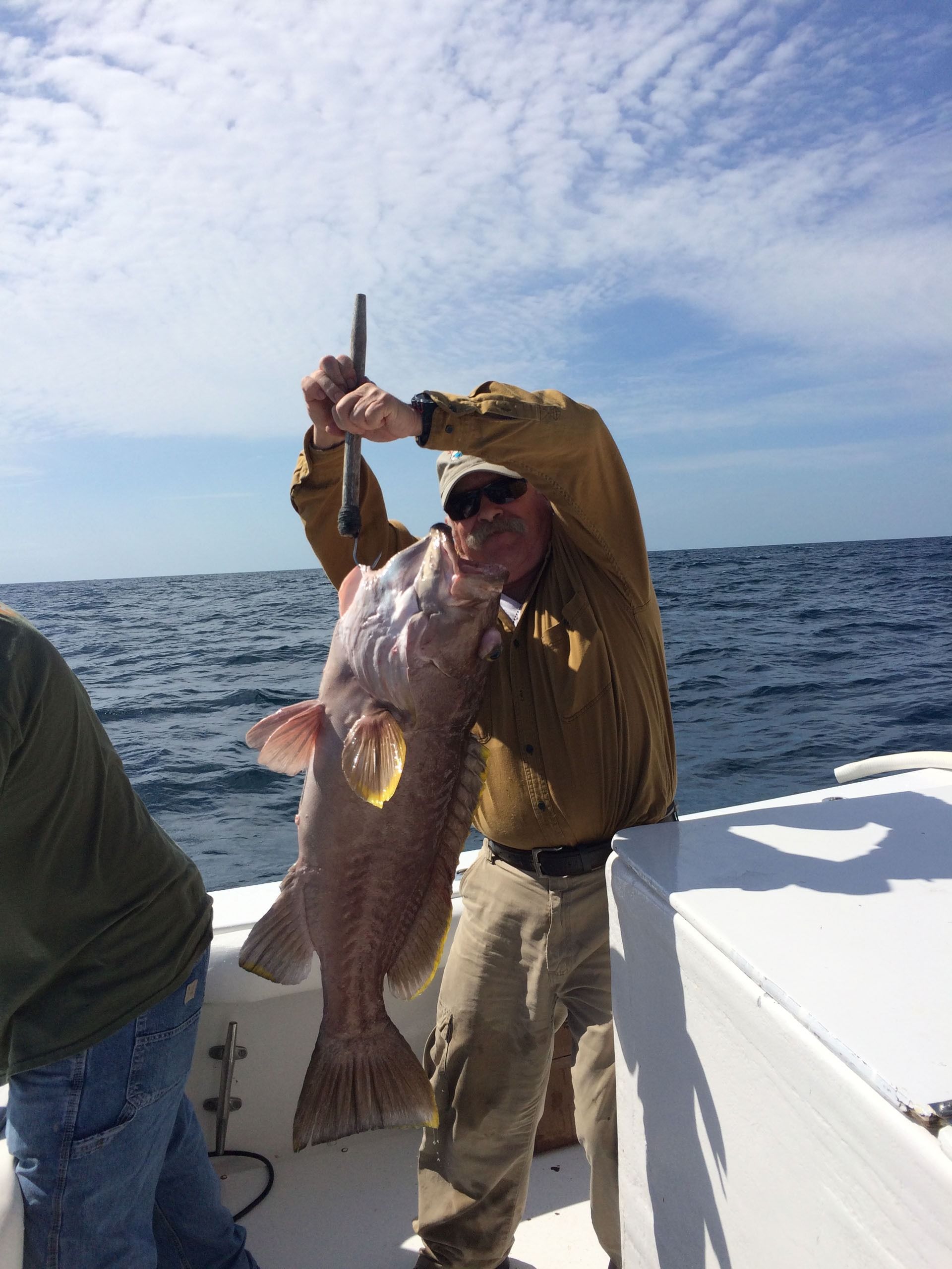 A person on a boat holding up a large fish by a metal tool, with the ocean and blue sky in the background.