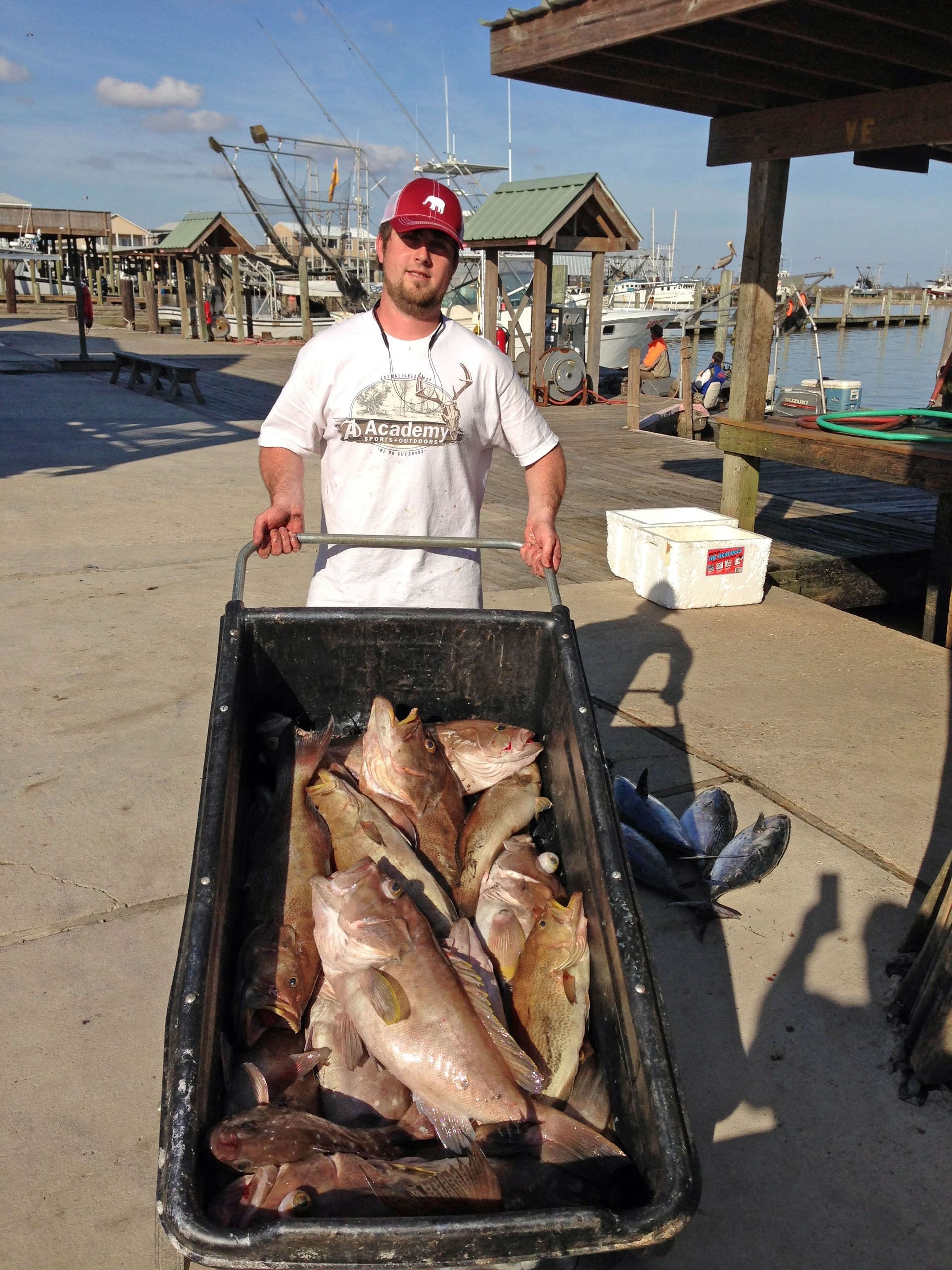 A person stands on a dock holding a large, black bin filled with freshly caught fish under a sunny, blue sky.