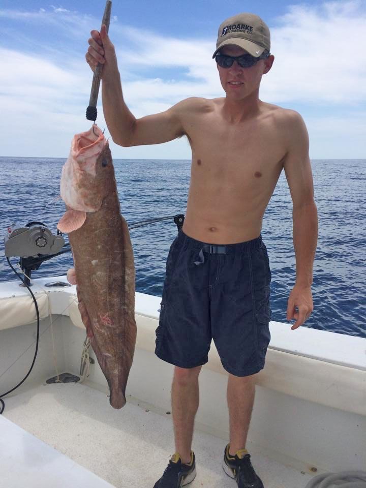 A person on a boat holding a large, brown fish hanging from a metal scale against an ocean background.