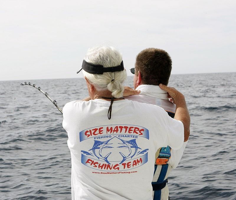 Two people on a boat, one assisting the other who is holding a fishing rod in the open ocean.