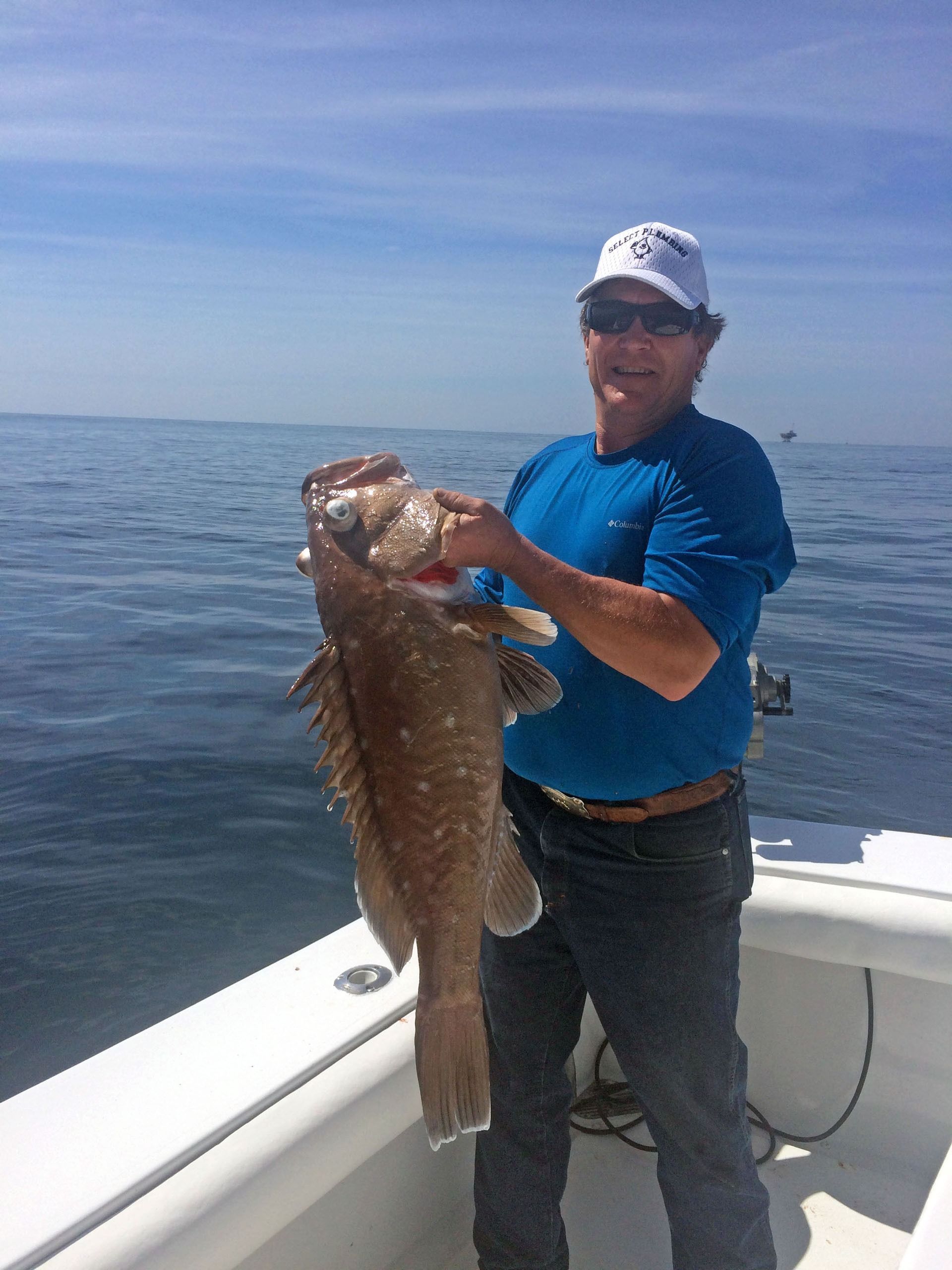 A person in a blue shirt and cap stands on a boat holding a large, speckled brown grouper fish over the open ocean.