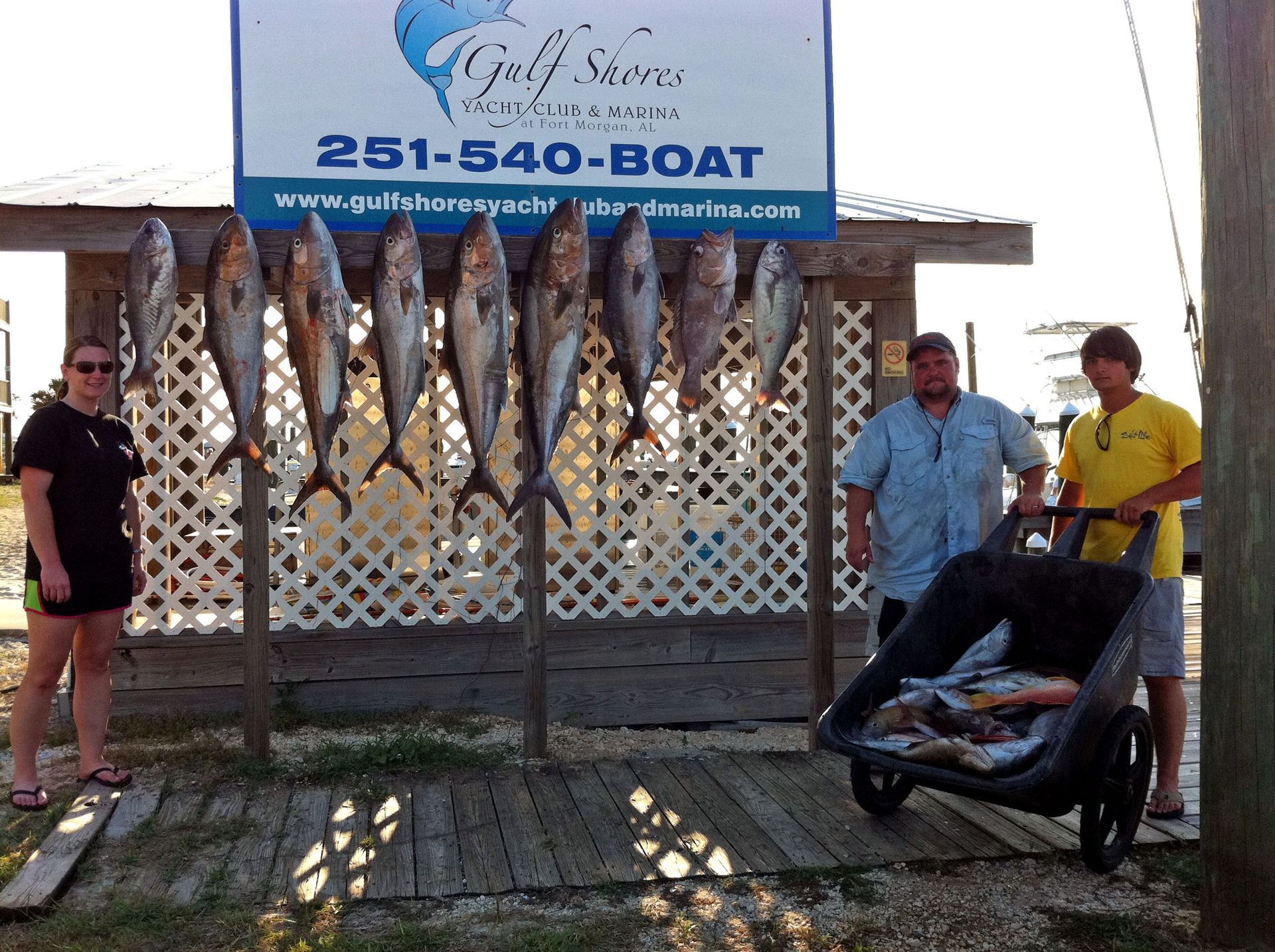 Three people pose with a catch of fish hanging from a rack at Gulf Shores Marina, with more fish in a cart.