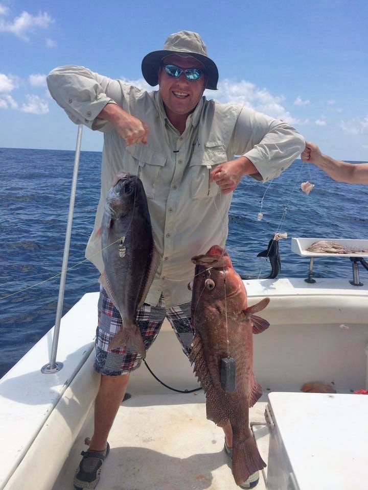 A person wearing a hat and sunglasses stands on a boat, smiling while holding two large fish, one in each hand.