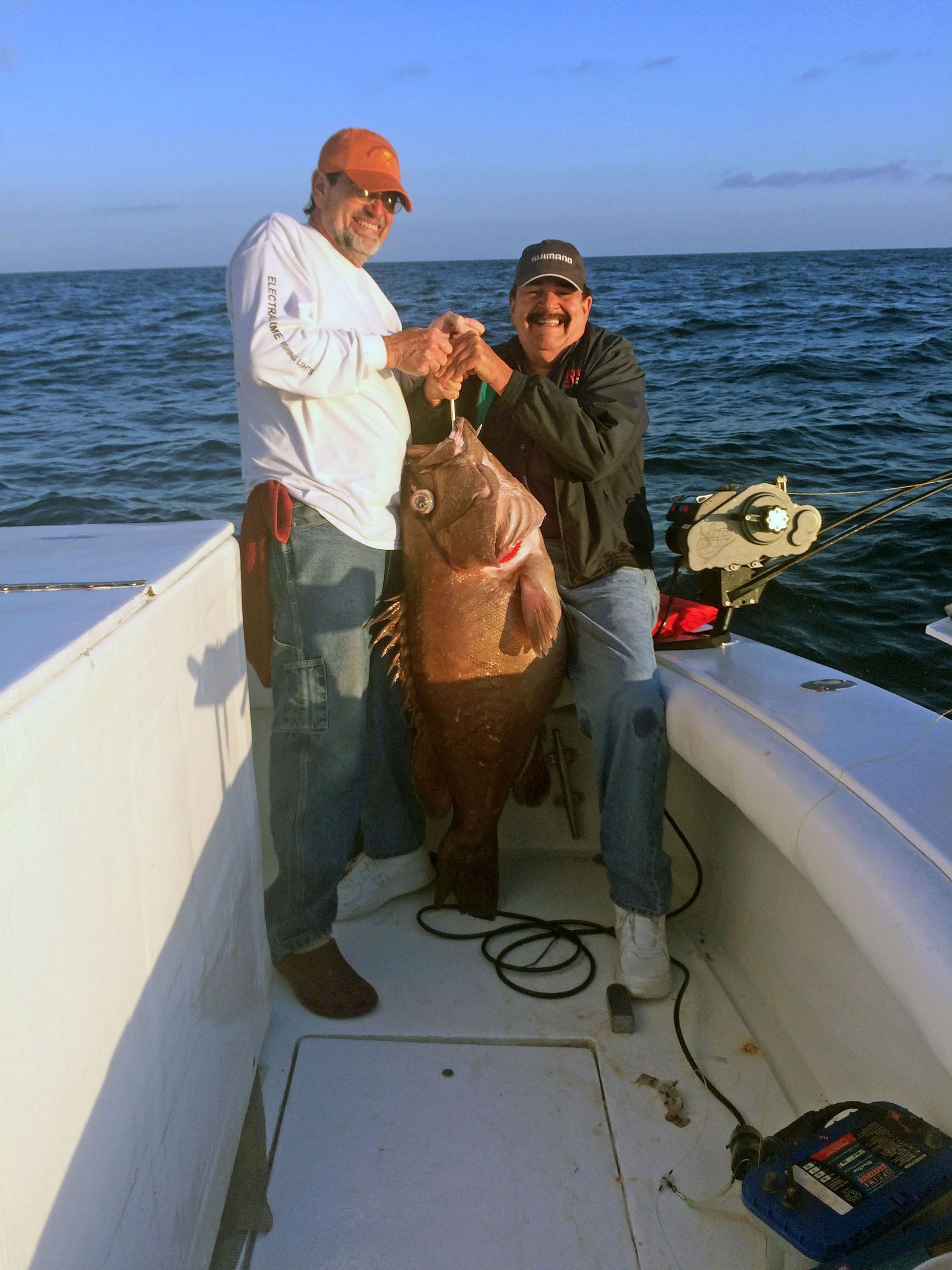 Two people stand on a boat deck, smiling and holding a large, brownish-gray grouper fish out of the water.