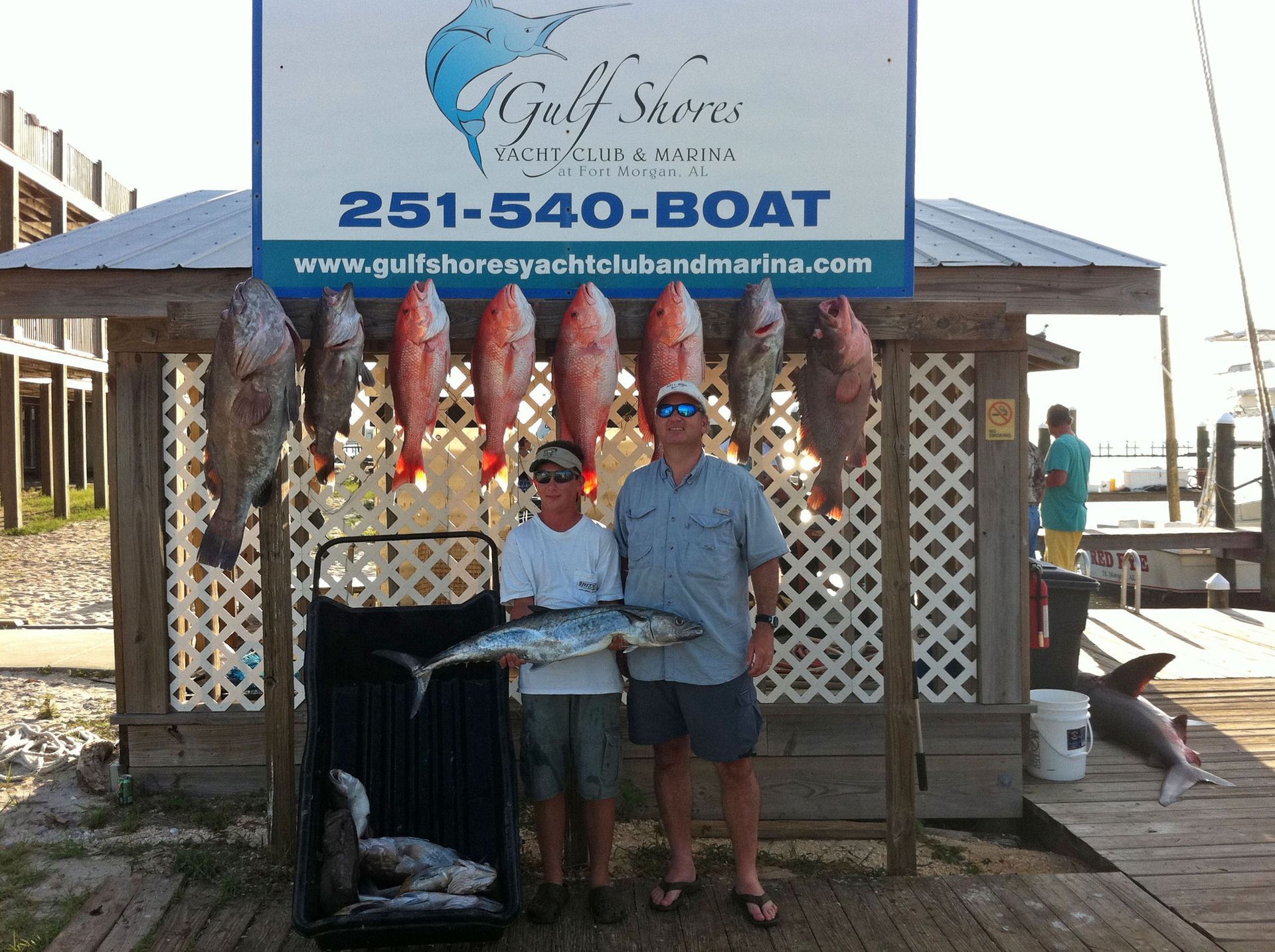 Two people standing in front of a marina sign with several fish hanging on a rack and a cooler of fish below.