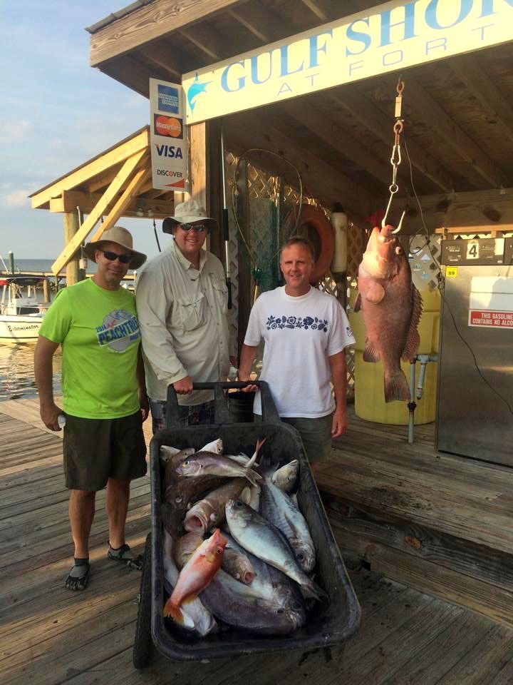 Three people stand on a dock at a fishing shop, posing behind a cart full of fish with a large fish hanging from a scale.