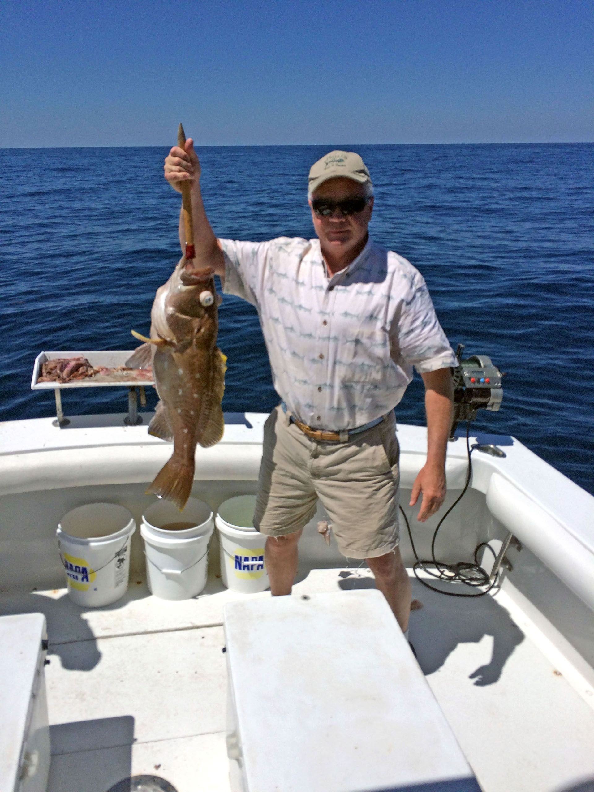 A person wearing a hat and sunglasses stands on a boat deck, holding up a freshly caught fish against the open ocean.
