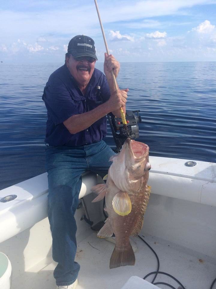 A person smiling while holding a large fish on a fishing rod on a boat at sea.