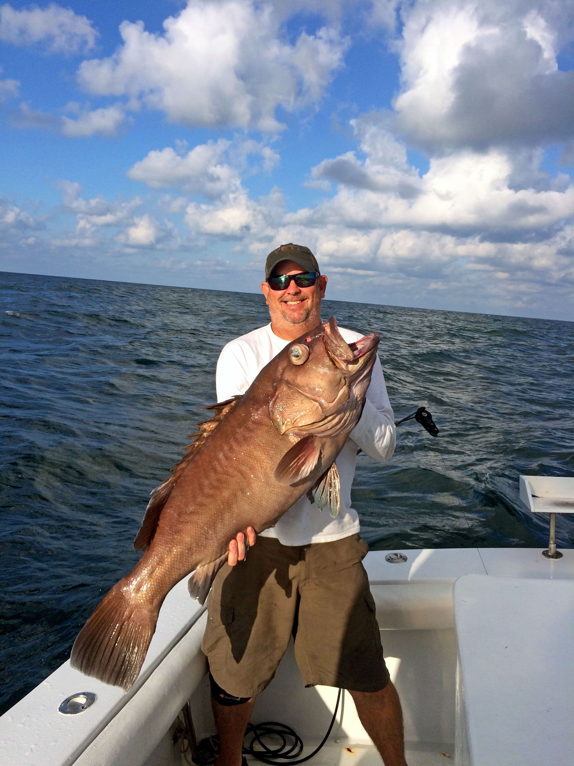 A person standing on a boat on the ocean, smiling while holding a large, brownish-gray grouper fish.