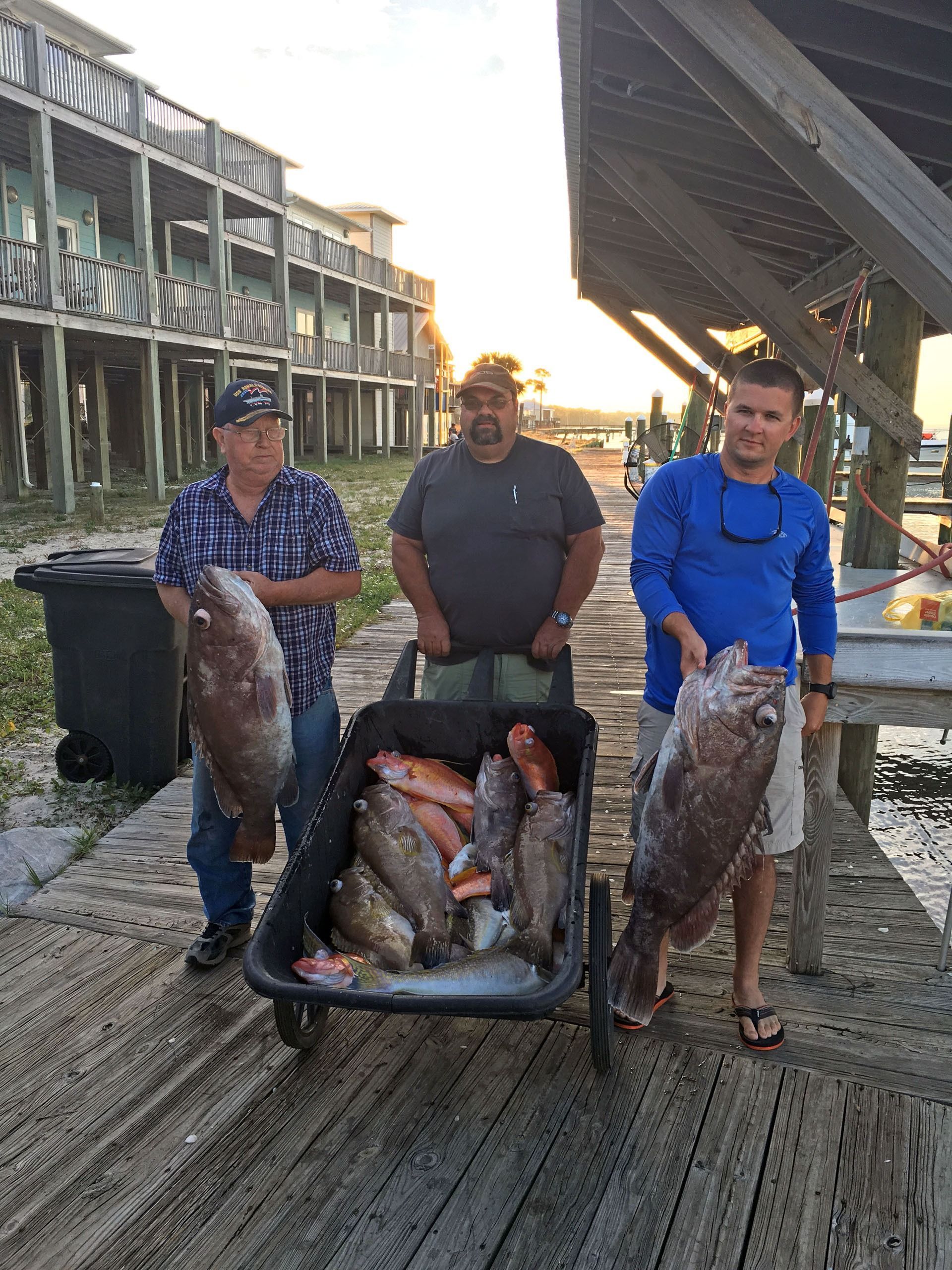Three people stand on a dock at sunset, holding large fish next to a wagon filled with a catch of fish.