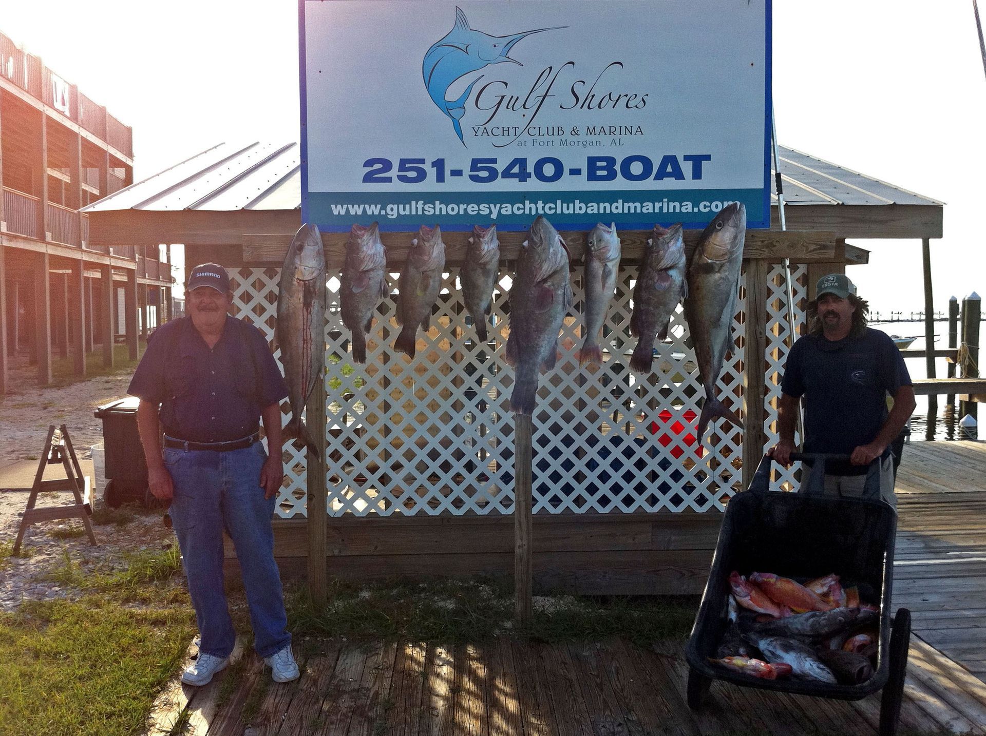 Two people stand beside a rack displaying a catch of fish in front of a Gulf Shores boat charter sign.