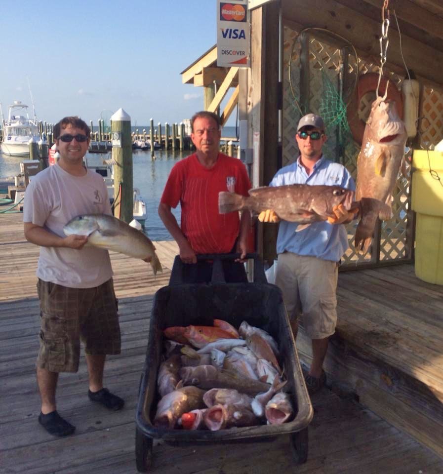 Three people stand on a wooden dock with a wheelbarrow full of freshly caught fish and hold individual large fish.