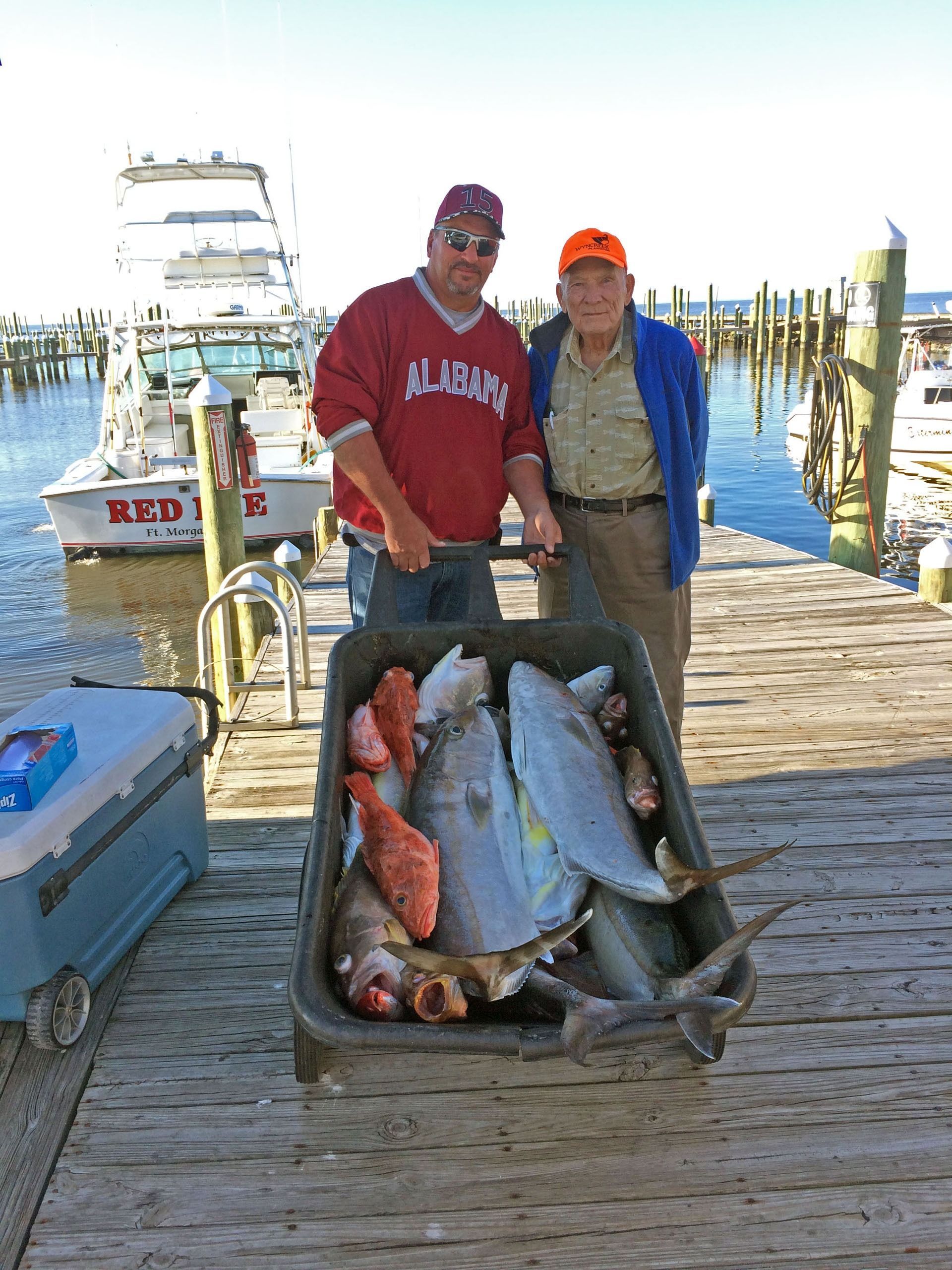 Two people standing on a dock behind a cart filled with large fish, with a boat in the background.