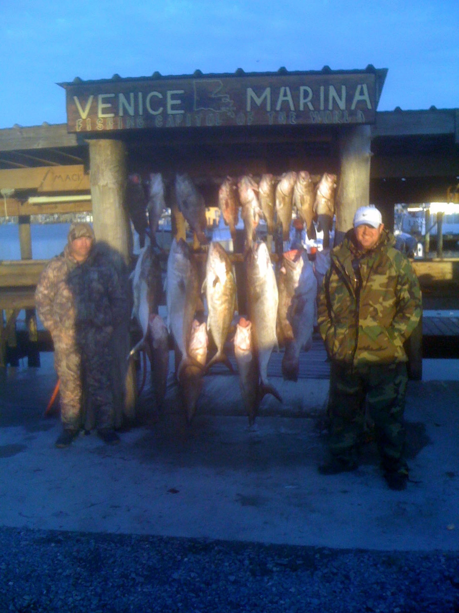 Two people in camo jackets stand by a large hanging catch of fish at Venice Marina.