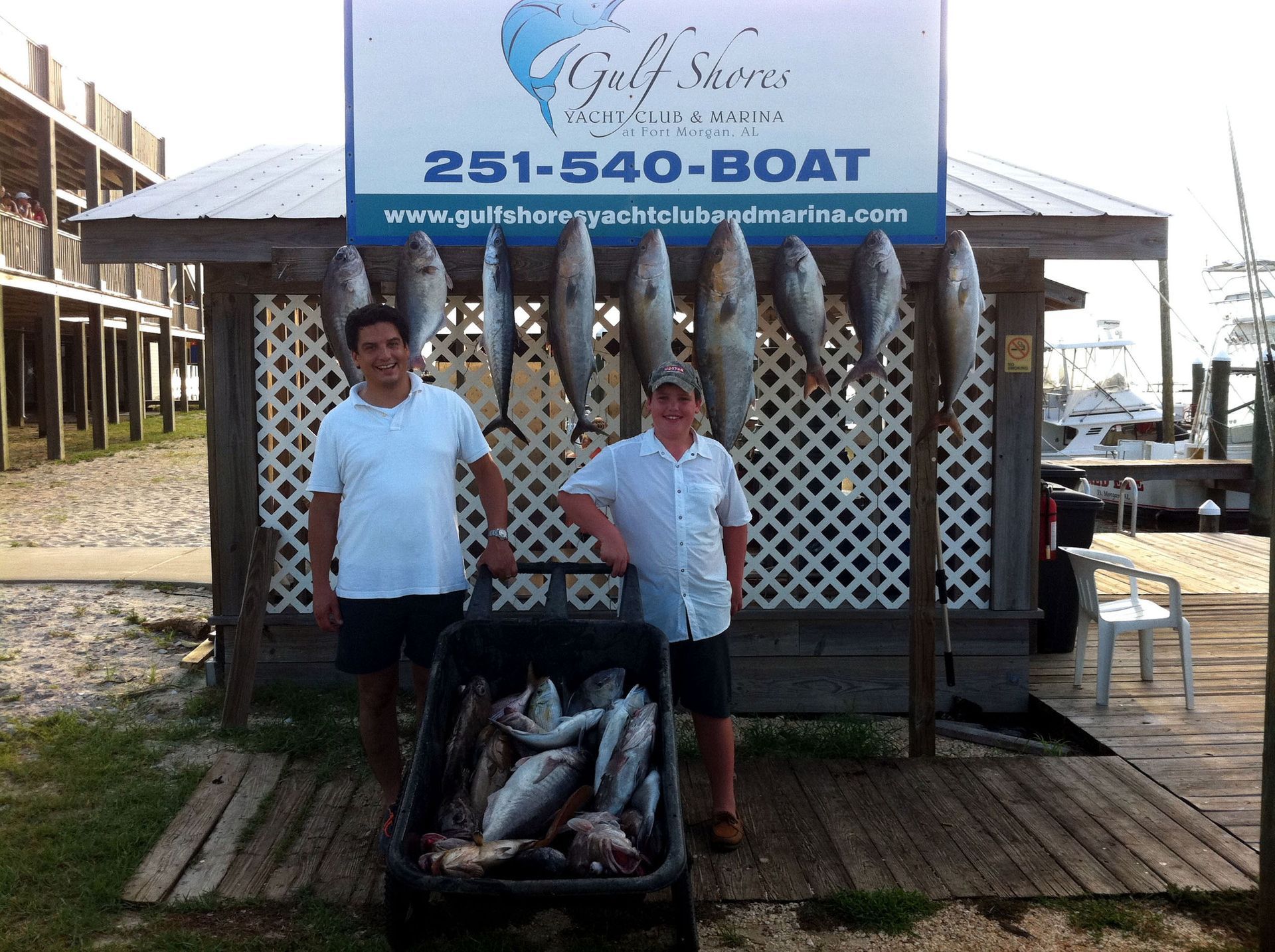 Two people stand before a marina sign and a rack of hanging fish, with a cooler of additional fish in the foreground.