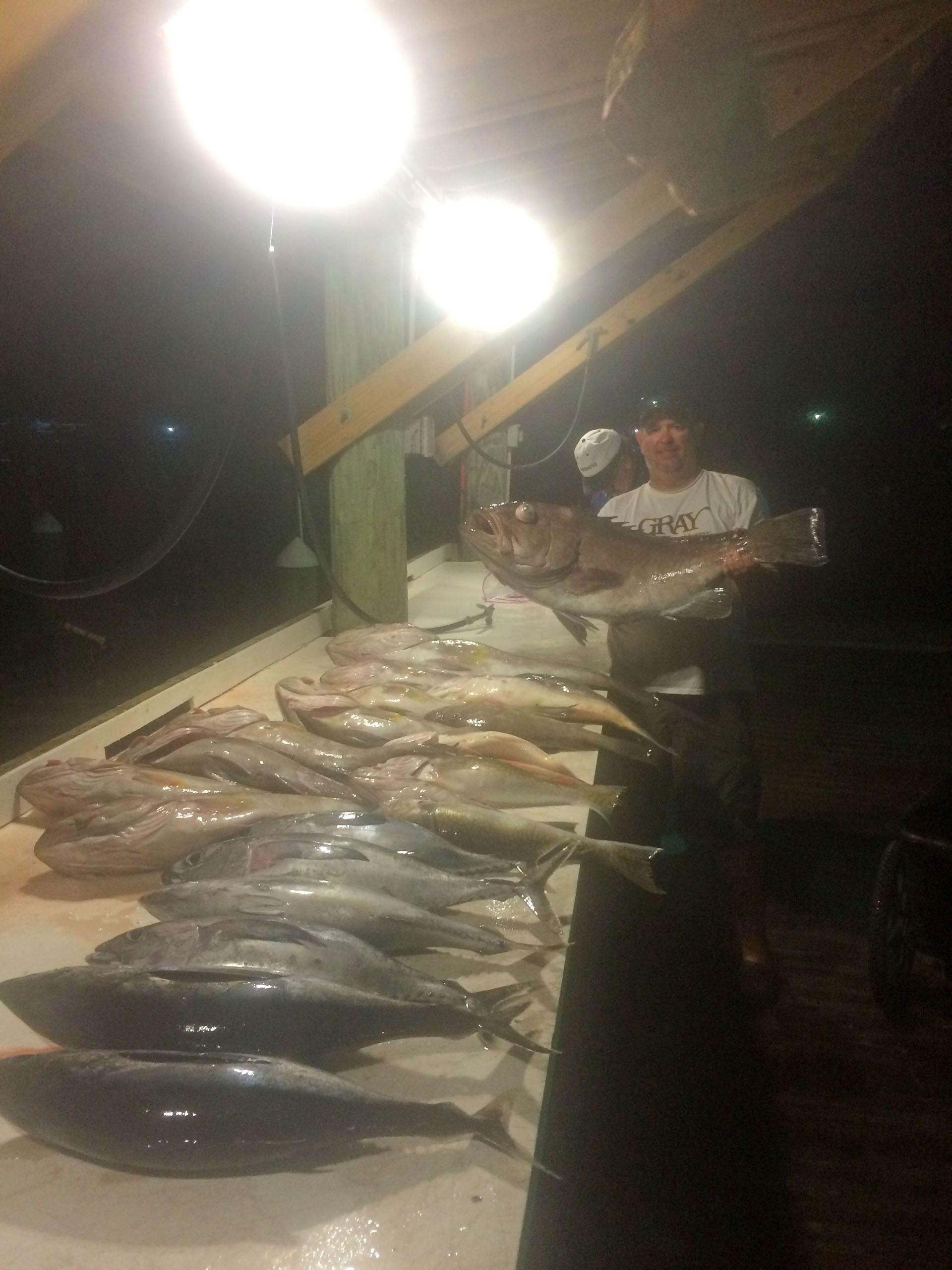 A person standing under bright lights at night, holding a large fish over a table covered in a catch of various fish.