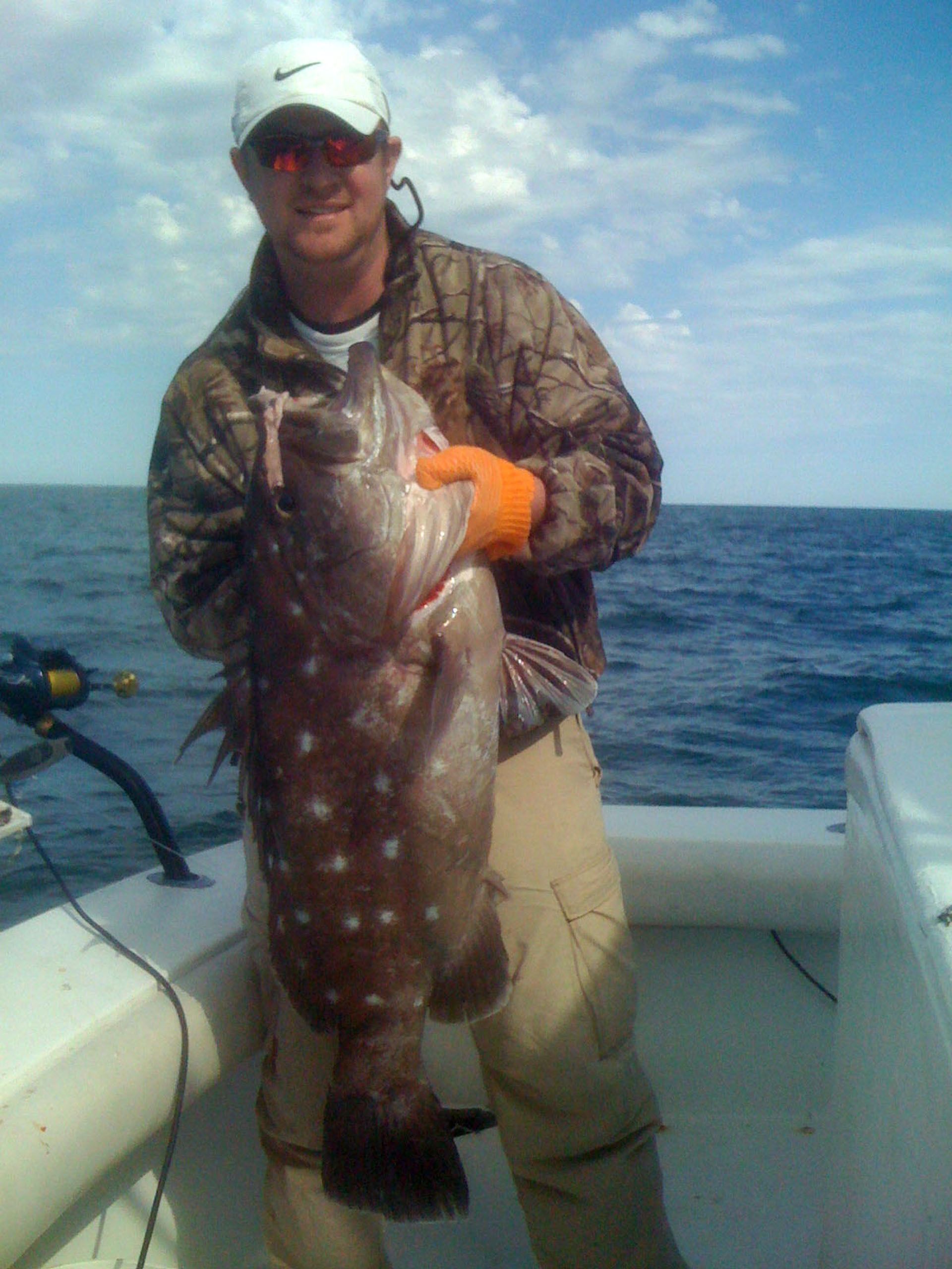 A person in a camouflage jacket and orange glove holds a large, brown, white-spotted grouper while on a boat at sea.