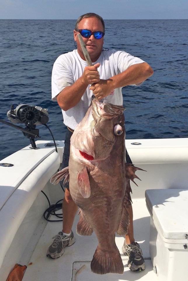 A person wearing sunglasses stands on a boat deck holding a large, caught fish by its mouth.